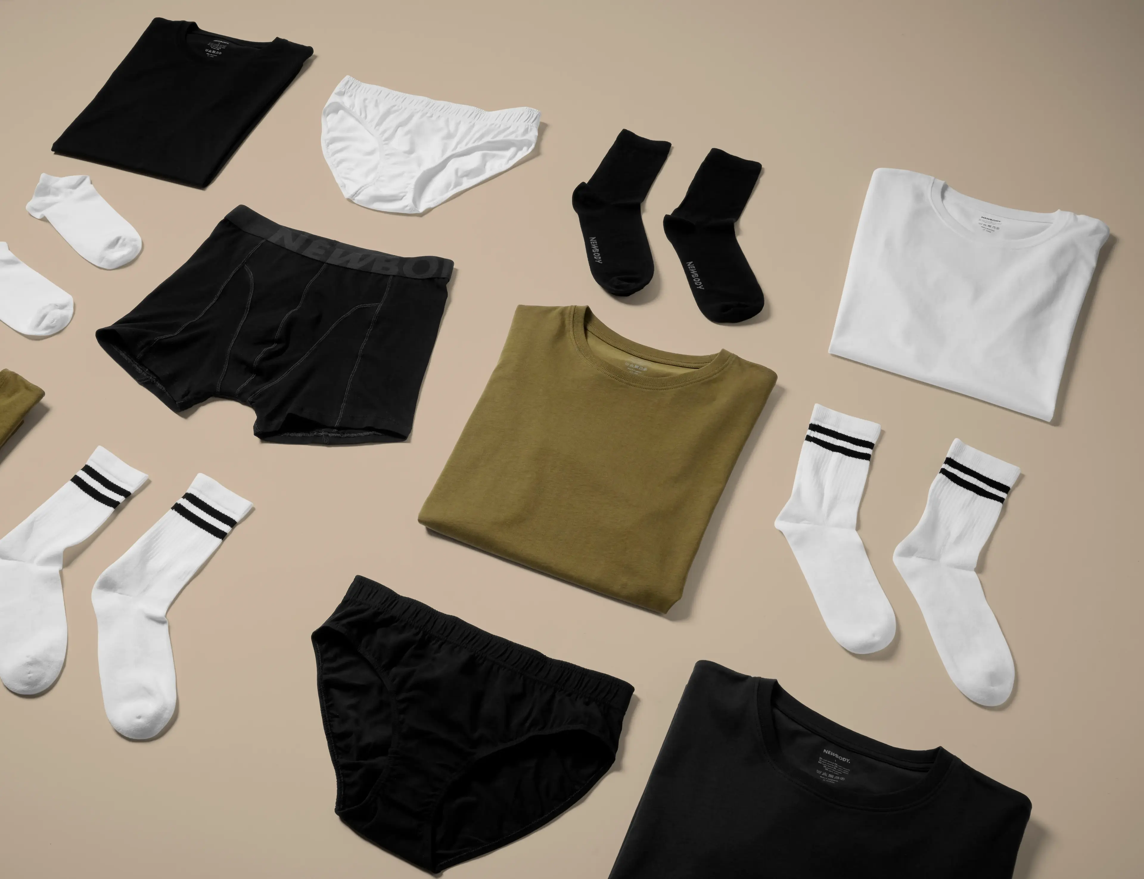 Olika plagg i neutrala färger, bland annat t-shirts, sockor och underkläder, snyggt upplagda på ett ljust underlag. Enkel design med fokus på basplagg för alla tillfällen.