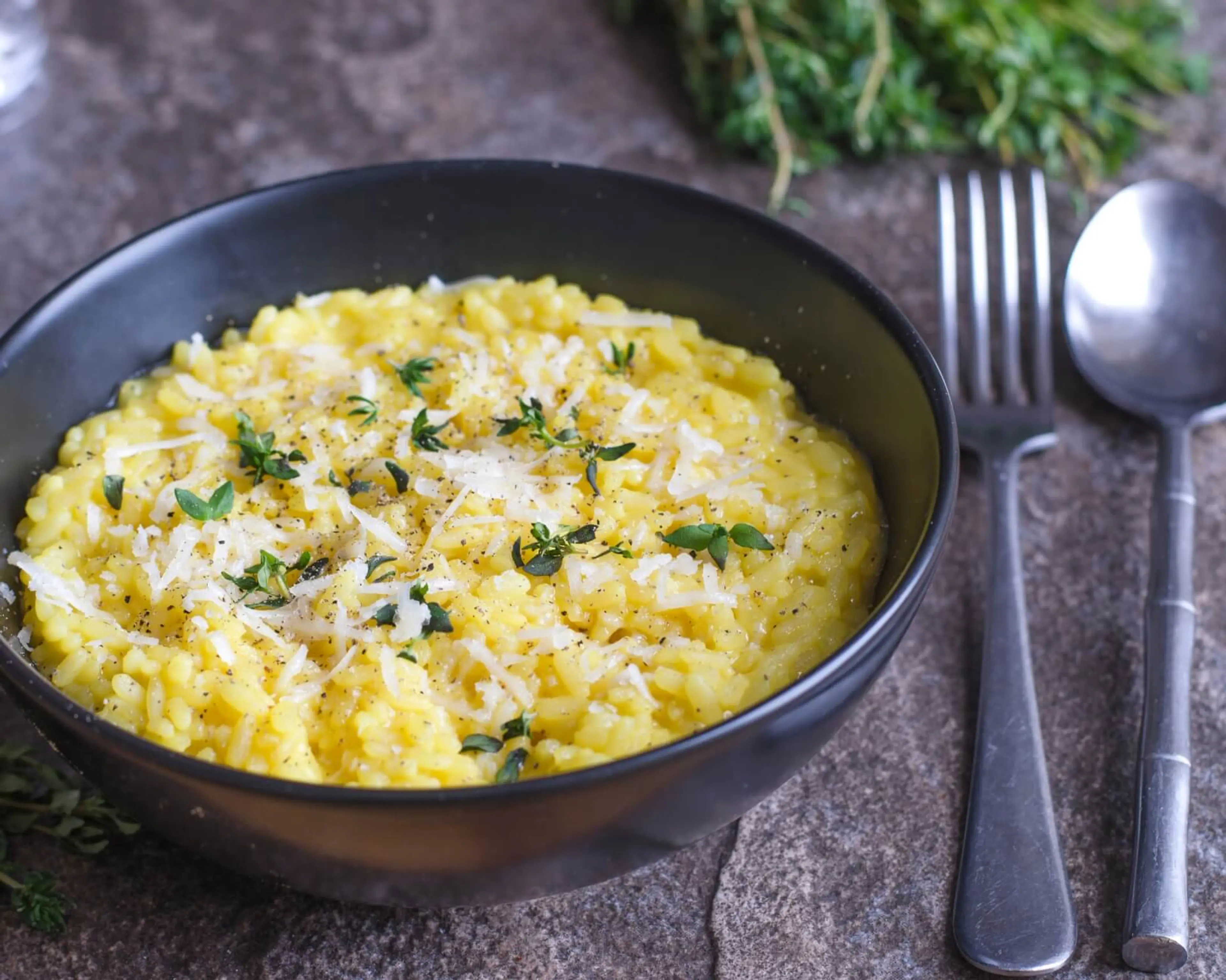 Krämig saffransrisotto toppad med riven parmesan och färsk timjan i en svart skål. Bredvid ligger en gaffel och en sked på en stenmönstrad yta.