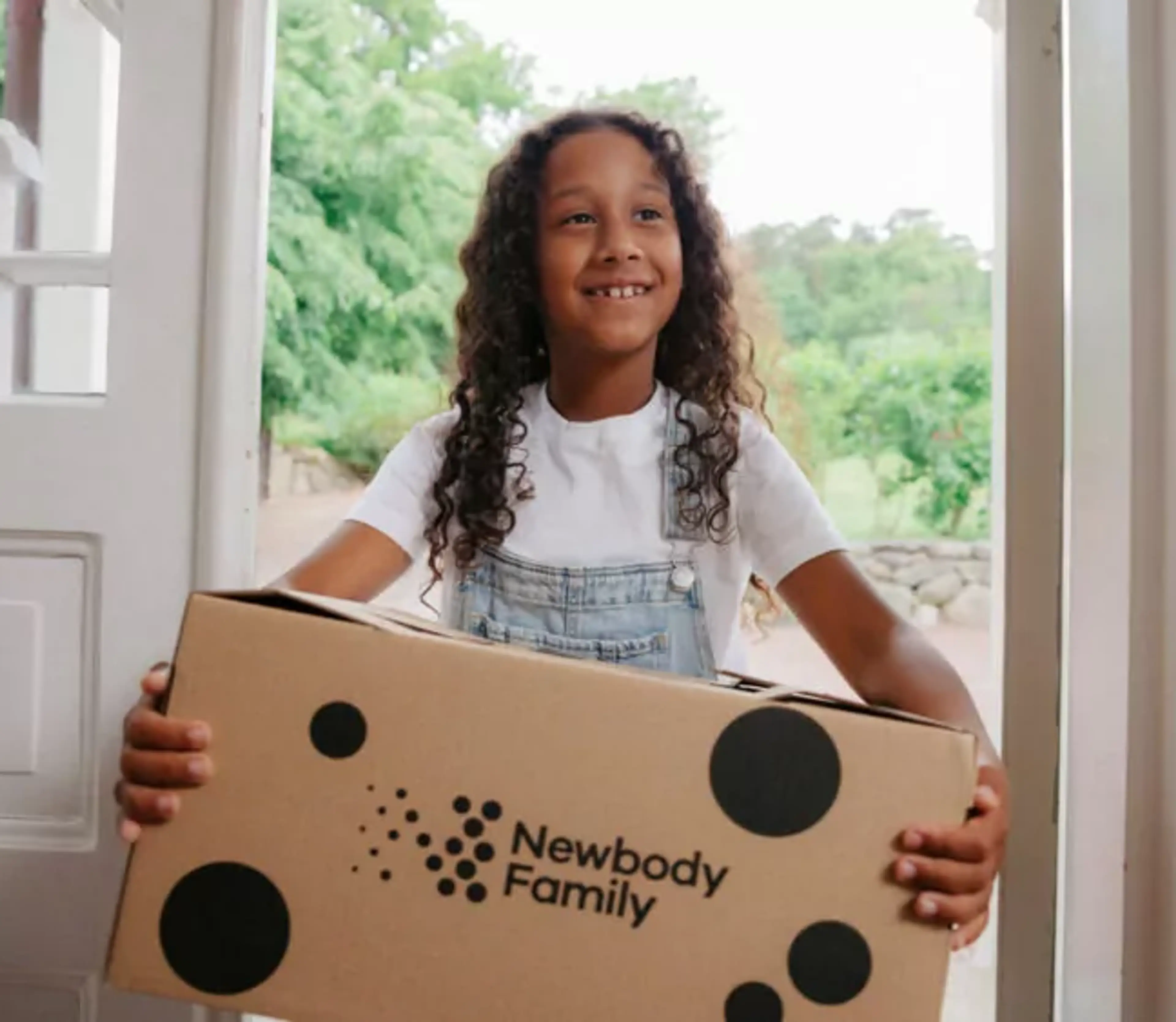 Ett barn med lockigt hår och ljus tröja bär en stor kartong med prickar och text. Det är dags för en leverans som precis har kommit hem.