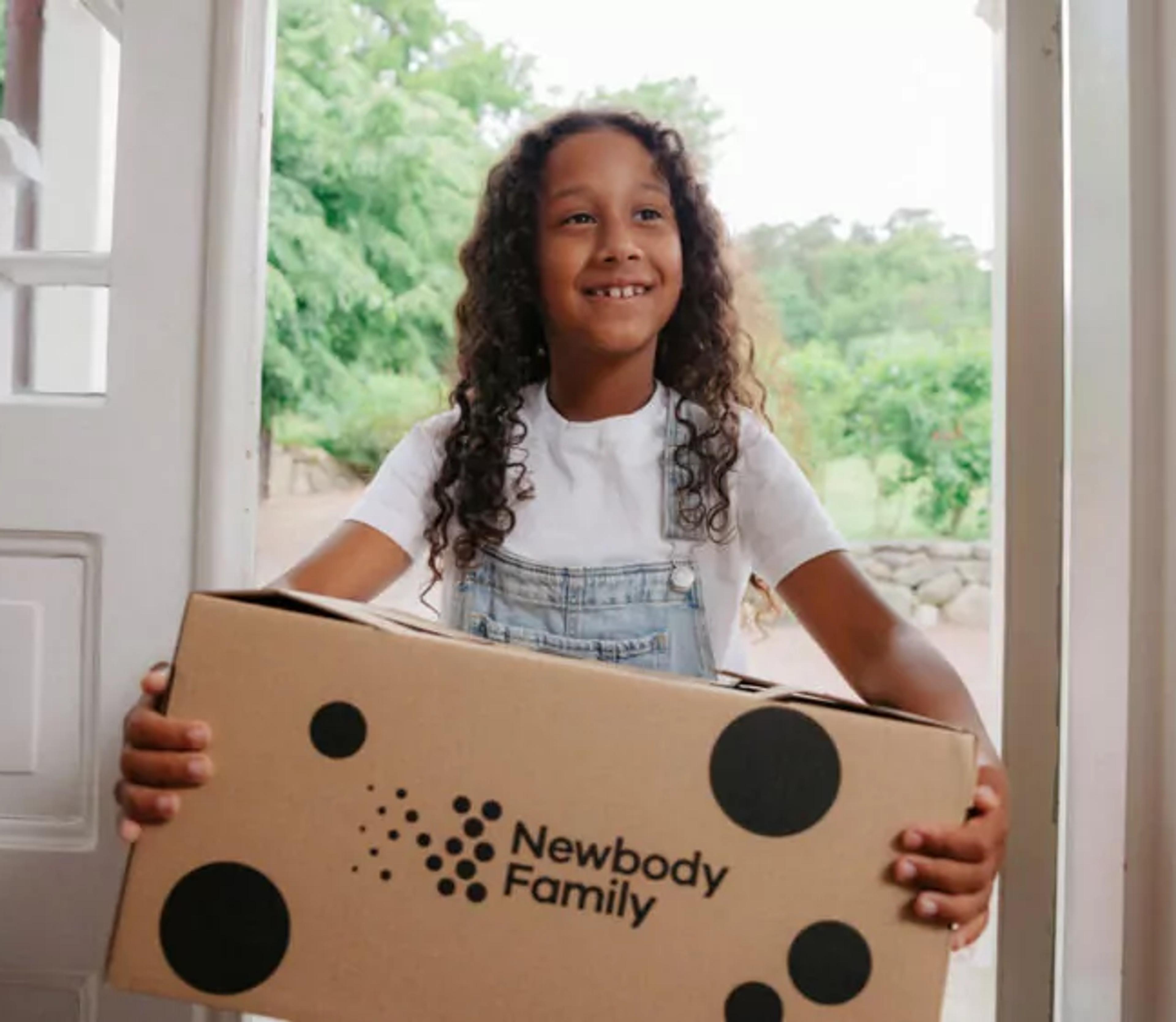 Ett barn med lockigt hår och ljus tröja bär en stor kartong med prickar och text. Det är dags för en leverans som precis har kommit hem.