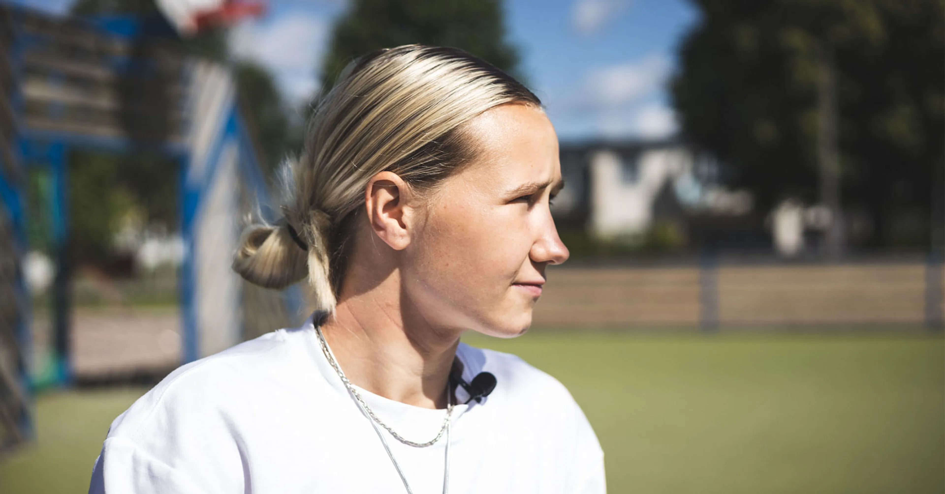 En person med blont hår uppsatt i en låg knut, iförd vit tröja, blickar mot något utanför bild. Bakgrunden är suddig med gröna och blå toner.