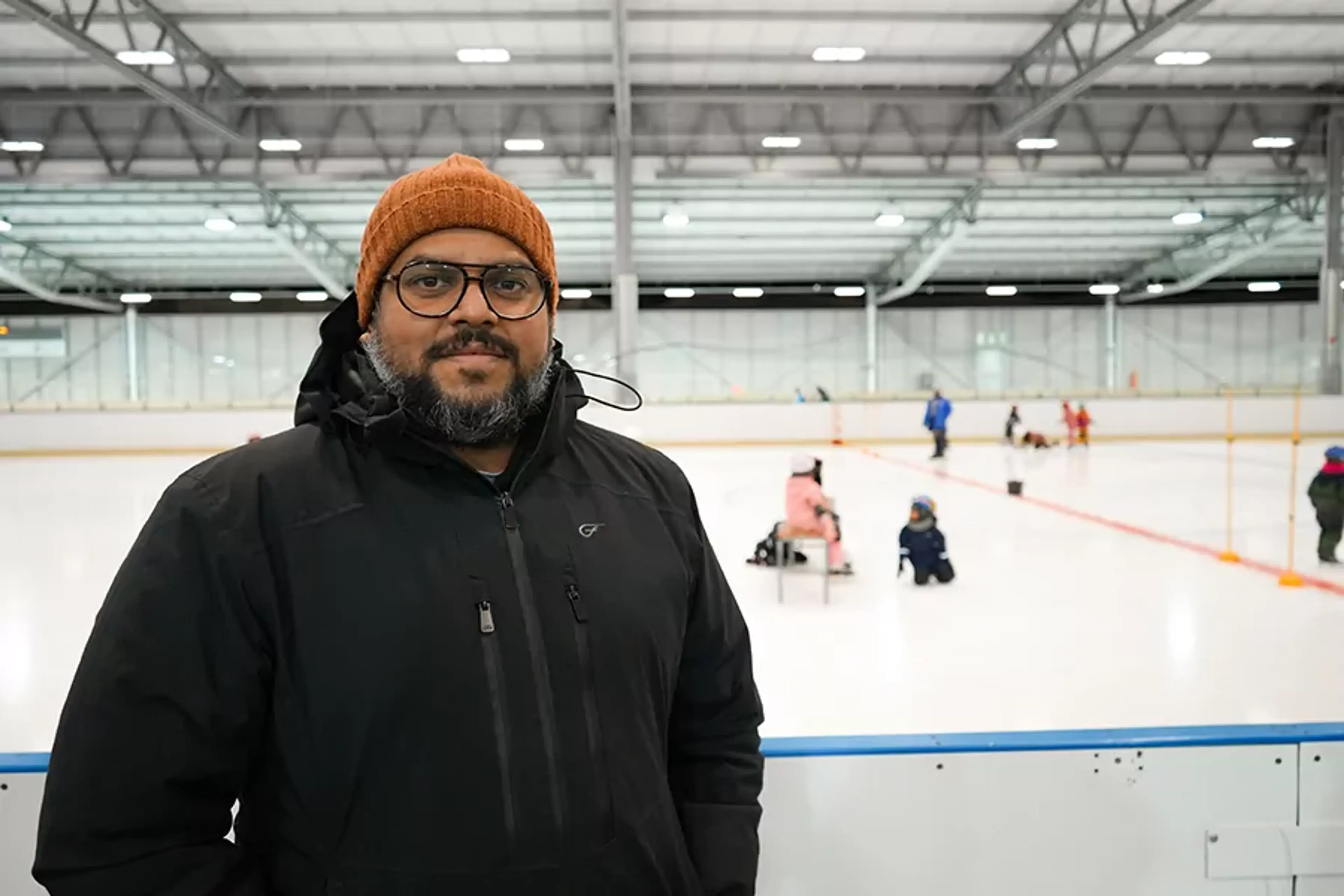 Man med orange mössa och svarta jacka står vid en ishockeyrink där barn på skridskor övar i bakgrunden. En inomhusanläggning med högt tak och konstgjord belysning.