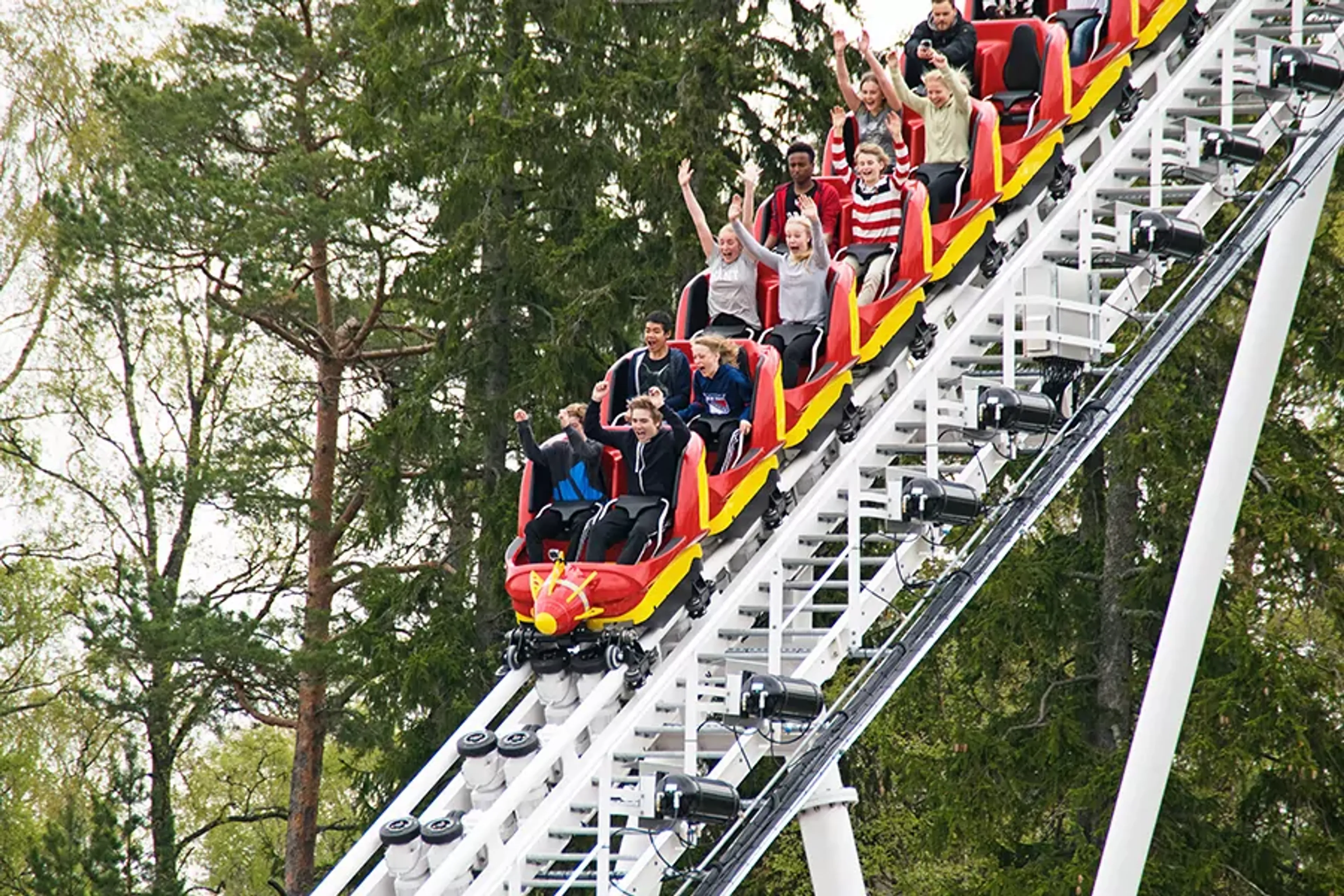 En grupp unga åker berg- och dalbana med armarna upp i luften på en nöjespark. Rullbanan går brant uppåt bland gröna träd.