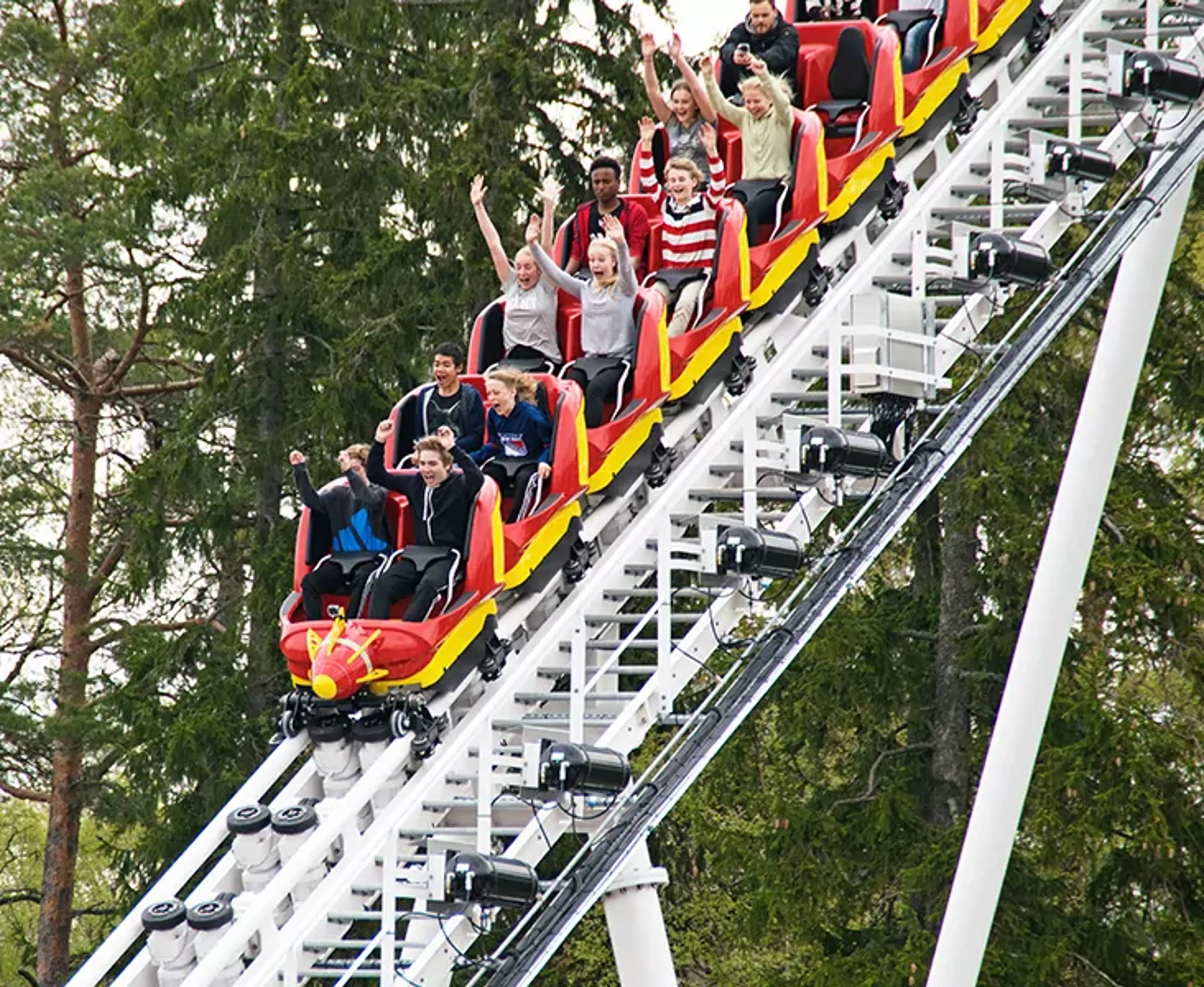 En grupp unga åker berg- och dalbana med armarna upp i luften på en nöjespark. Rullbanan går brant uppåt bland gröna träd.