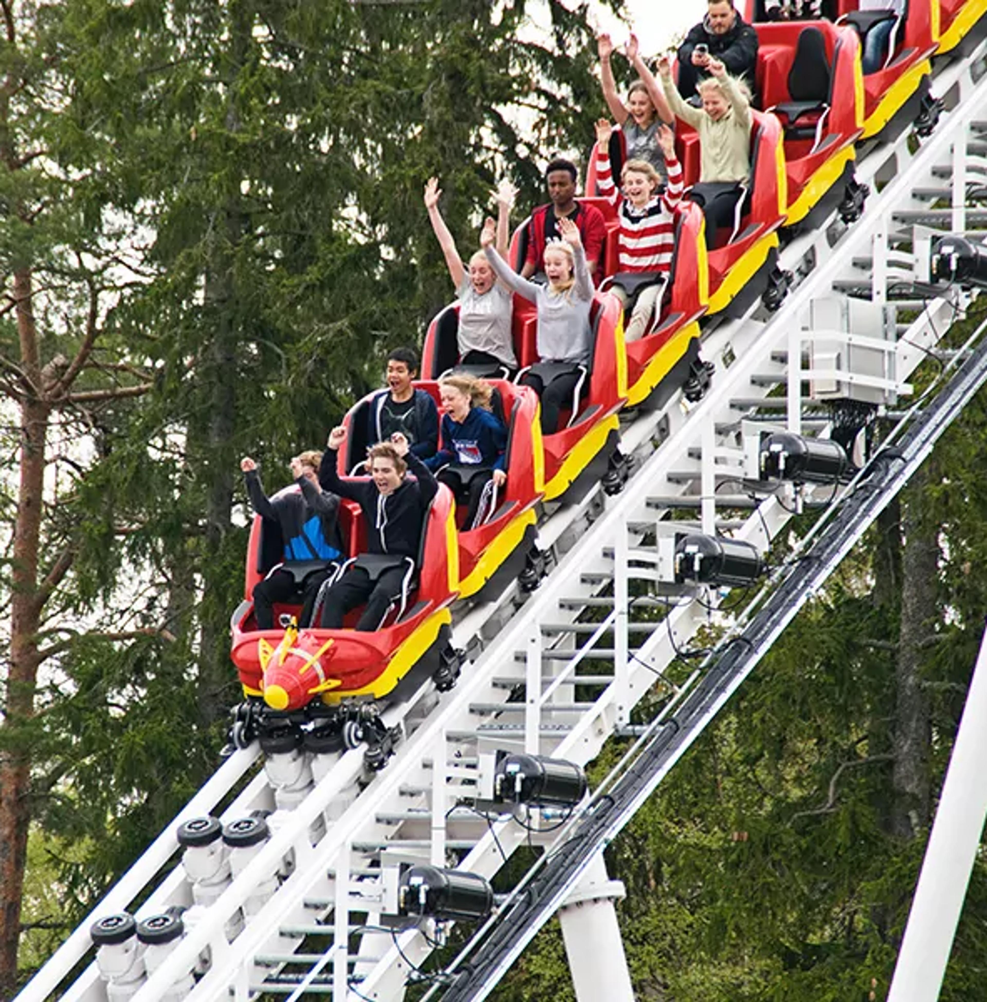 En grupp unga åker berg- och dalbana med armarna upp i luften på en nöjespark. Rullbanan går brant uppåt bland gröna träd.