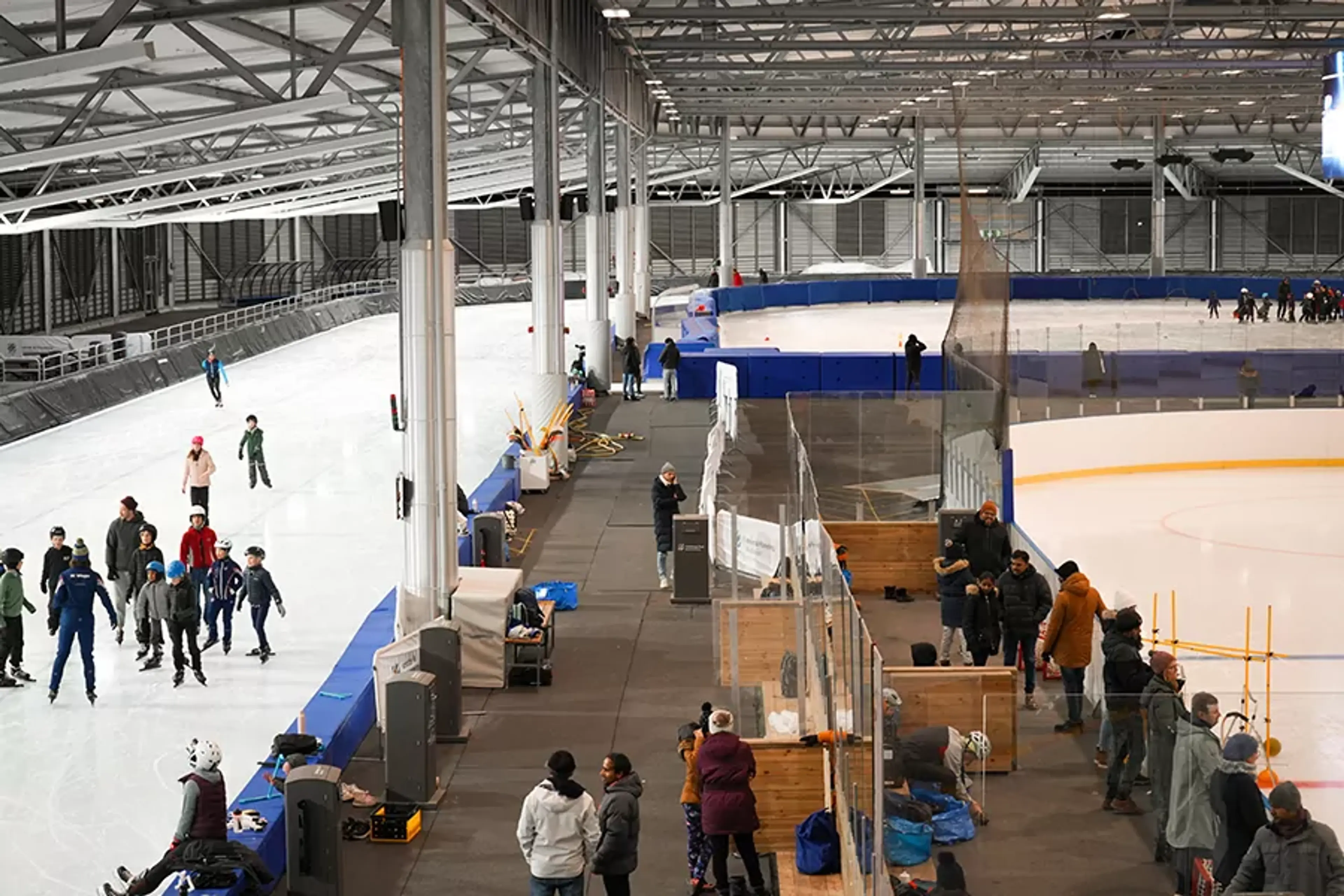En inomhushall med flera personer som åker skridskor på isbanan och andra som står och pratar vid sidan av. Den rymliga miljön är fylld med naturligt ljus från stora takfönster.