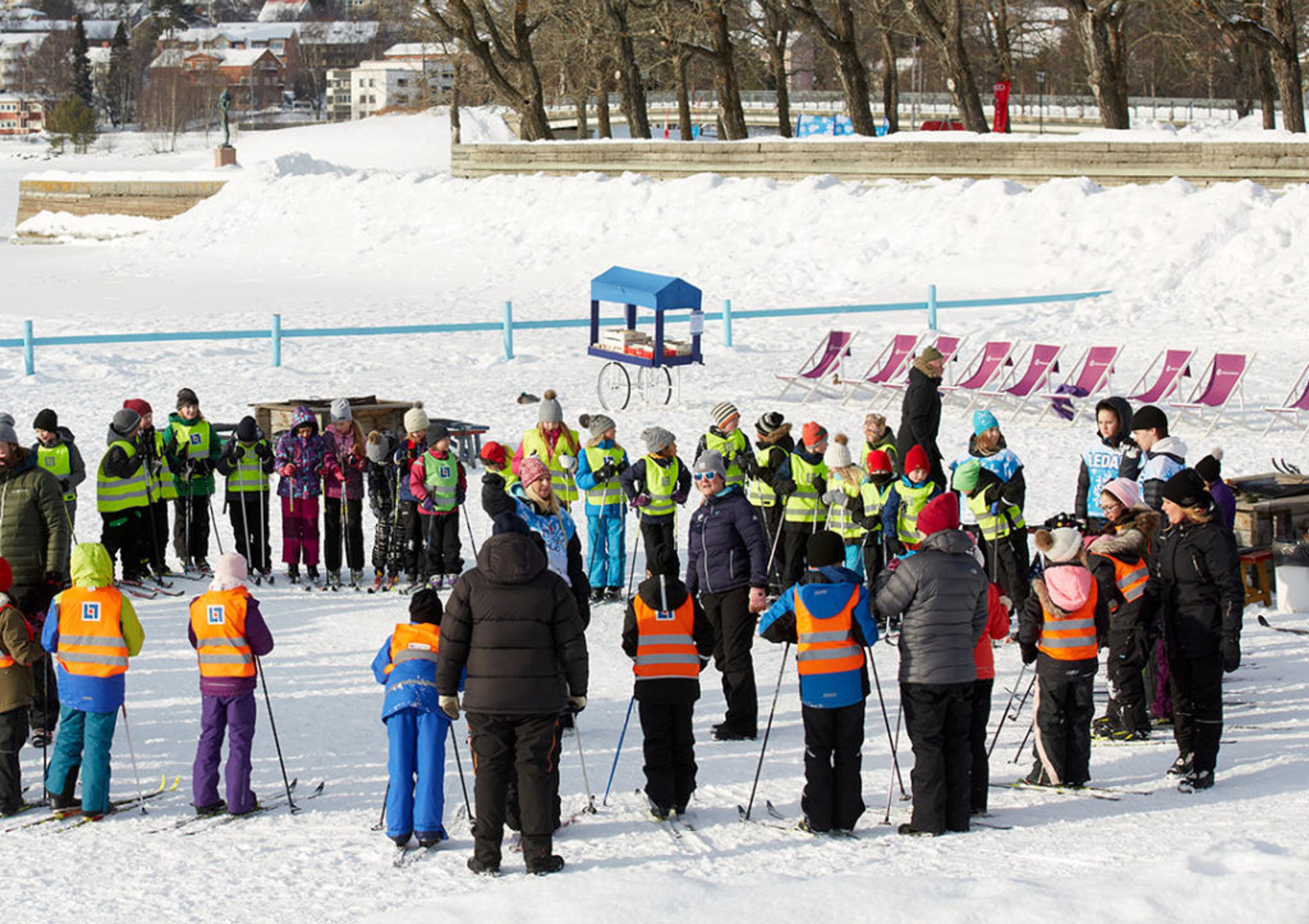 Barn och vuxna i färgglada vinterkläder samlade i en stor cirkel på snön under en skidlektion. I bakgrunden syns träd, hus och en skidbacke.