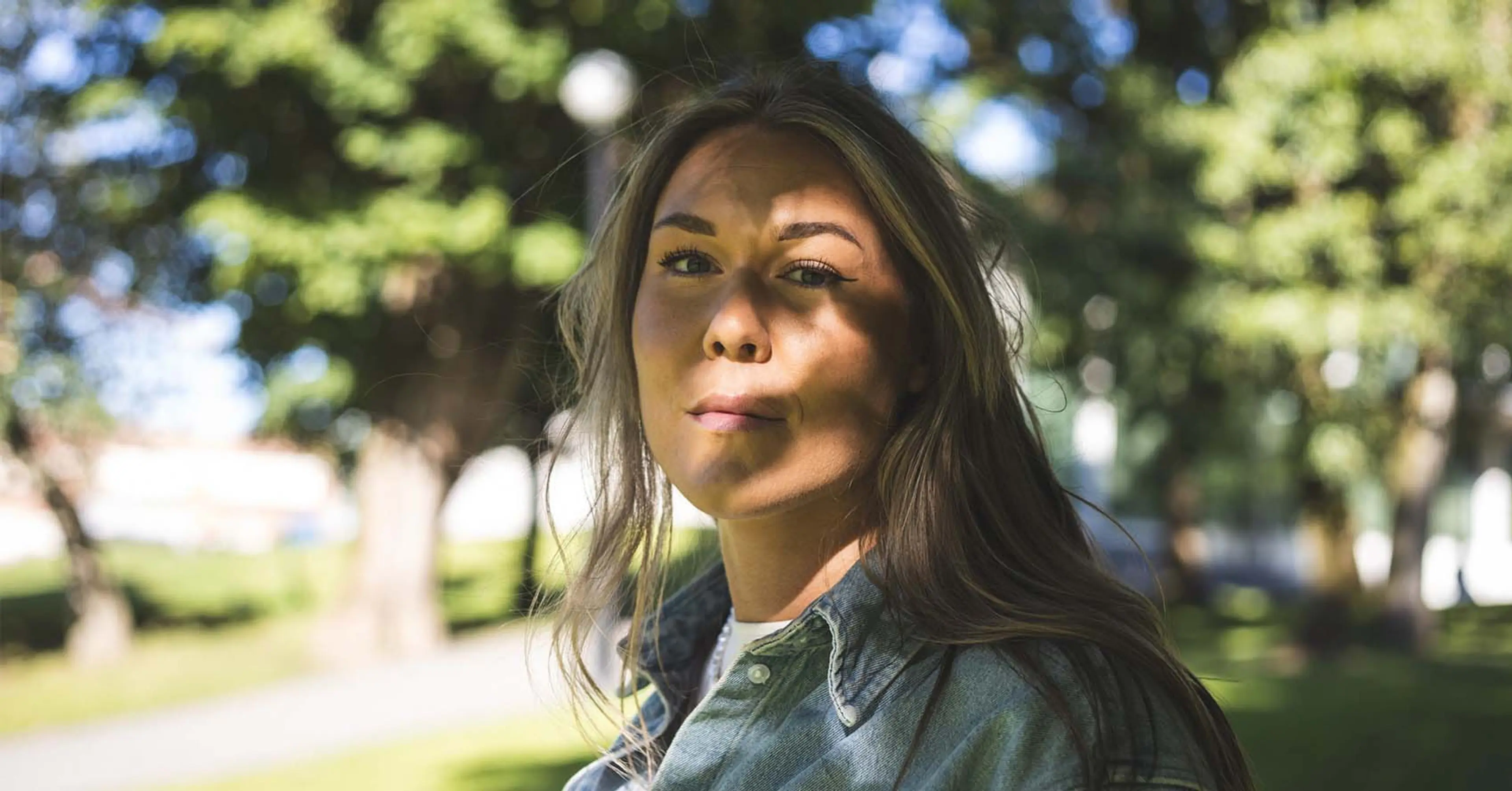 En person med långt hår och jeansskjorta står utomhus med gröna träd i bakgrunden. Solens strålar skapar skuggmönster över hennes ansikte.