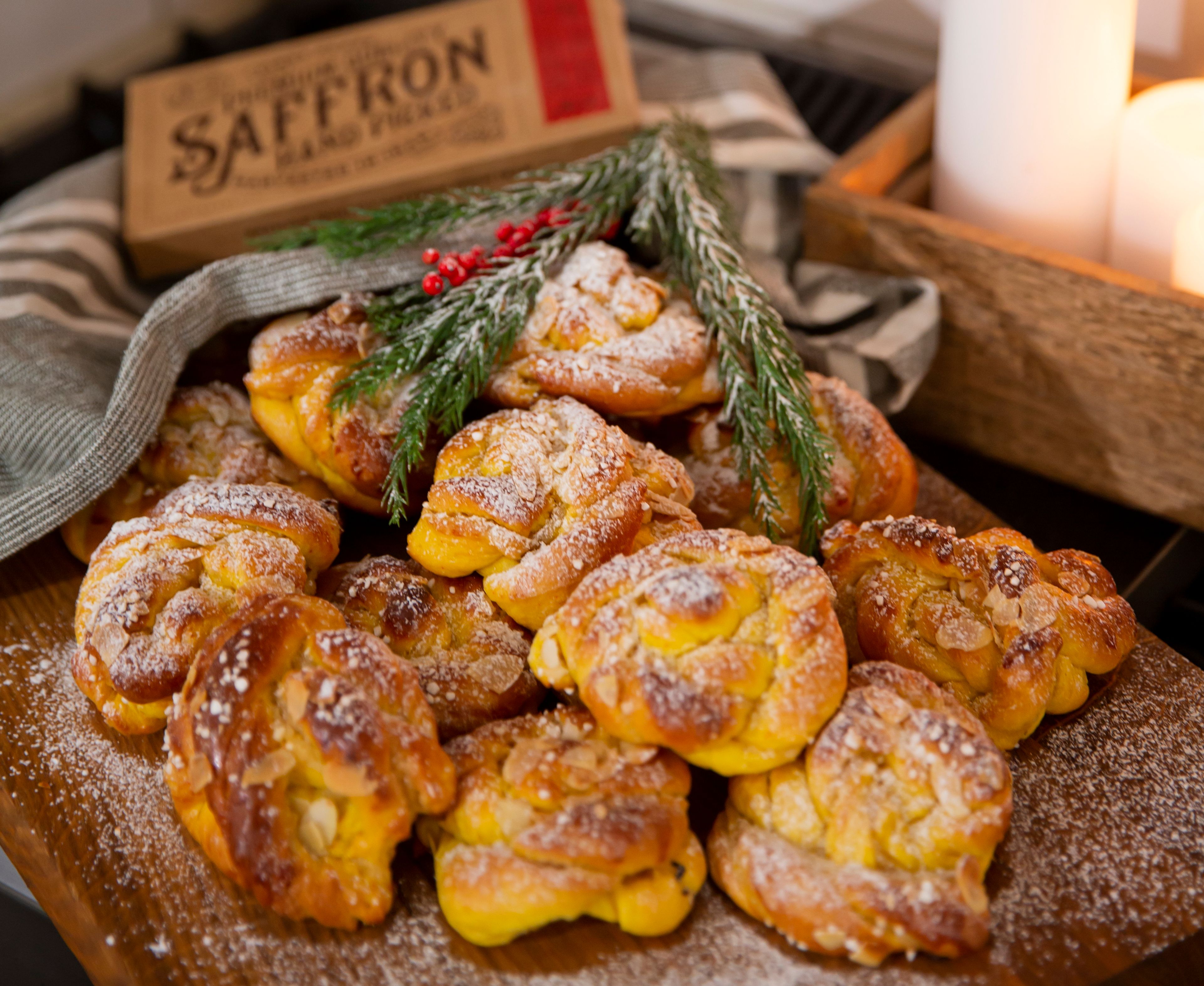 Saftiga saffransbullar med mandelspån och pudrat florsocker, uppdukade på en träbricka tillsammans med en ask saffran och vintergrönt. Tre tända vita blockljus ger ett varmt sken i bakgrunden.