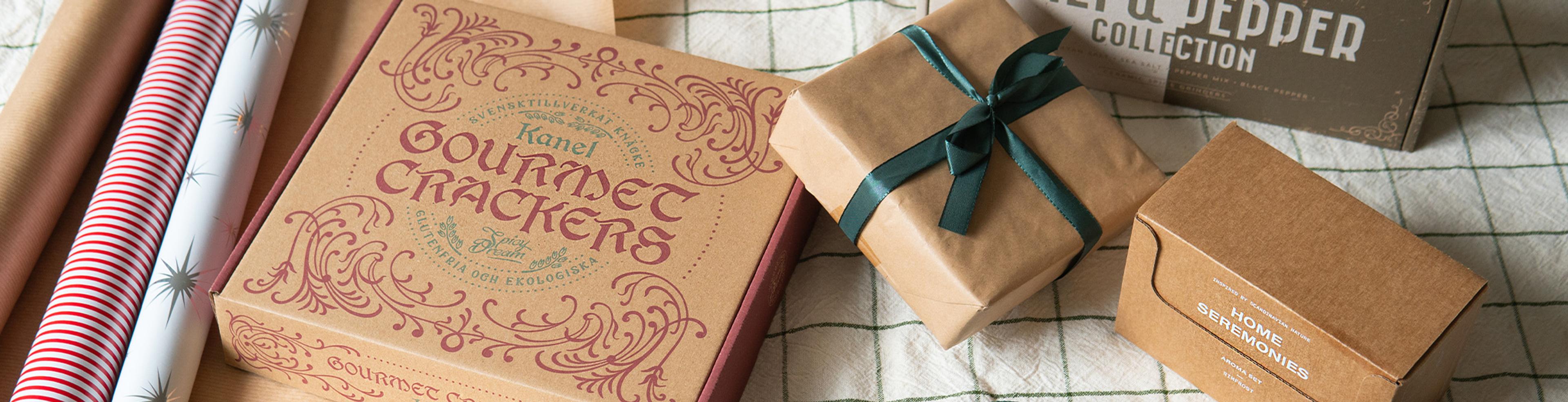 Flera inslagna paket med brunt papper och ett grönt band tillsammans med förpackningar av gourmetkex och kryddor på en rutig duk. Rullarna med presentpapper i olika mönster ligger bredvid paketen.