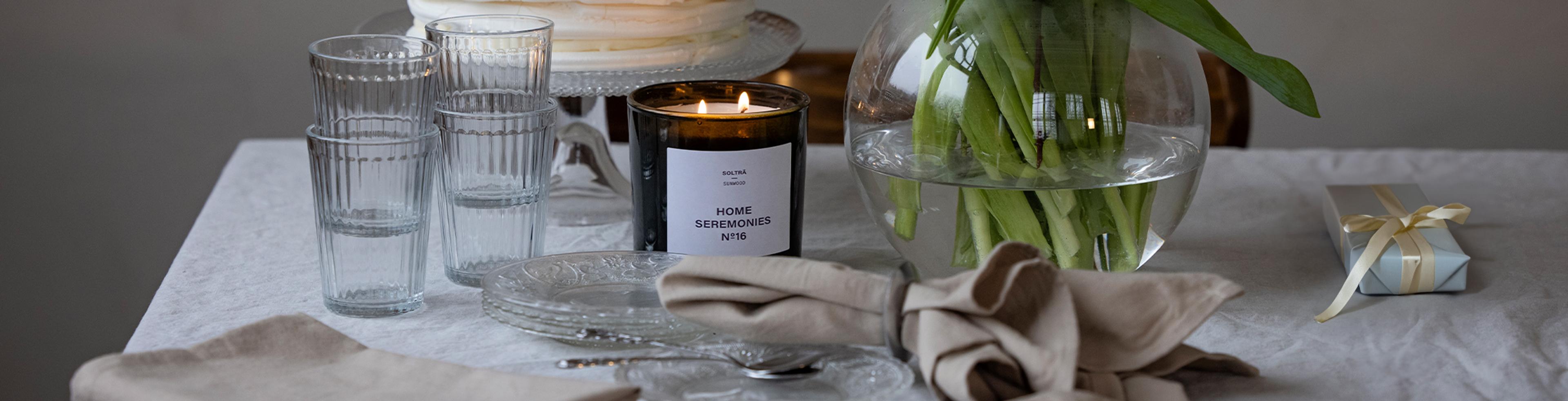 Ett bord med staplade glas, en tänd svart doftljusburk med texten "Home Seremonies", en vas med gröna växtstjälkar och en liten presentpaket med cremefärgat band. Dukarna är i ljusa, naturf