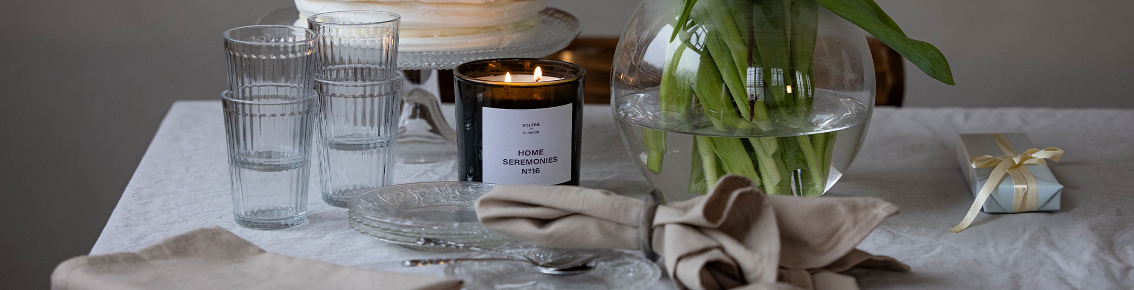 Ett bord med staplade glas, en tänd svart doftljusburk med texten "Home Seremonies", en vas med gröna växtstjälkar och en liten presentpaket med cremefärgat band. Dukarna är i ljusa, naturf