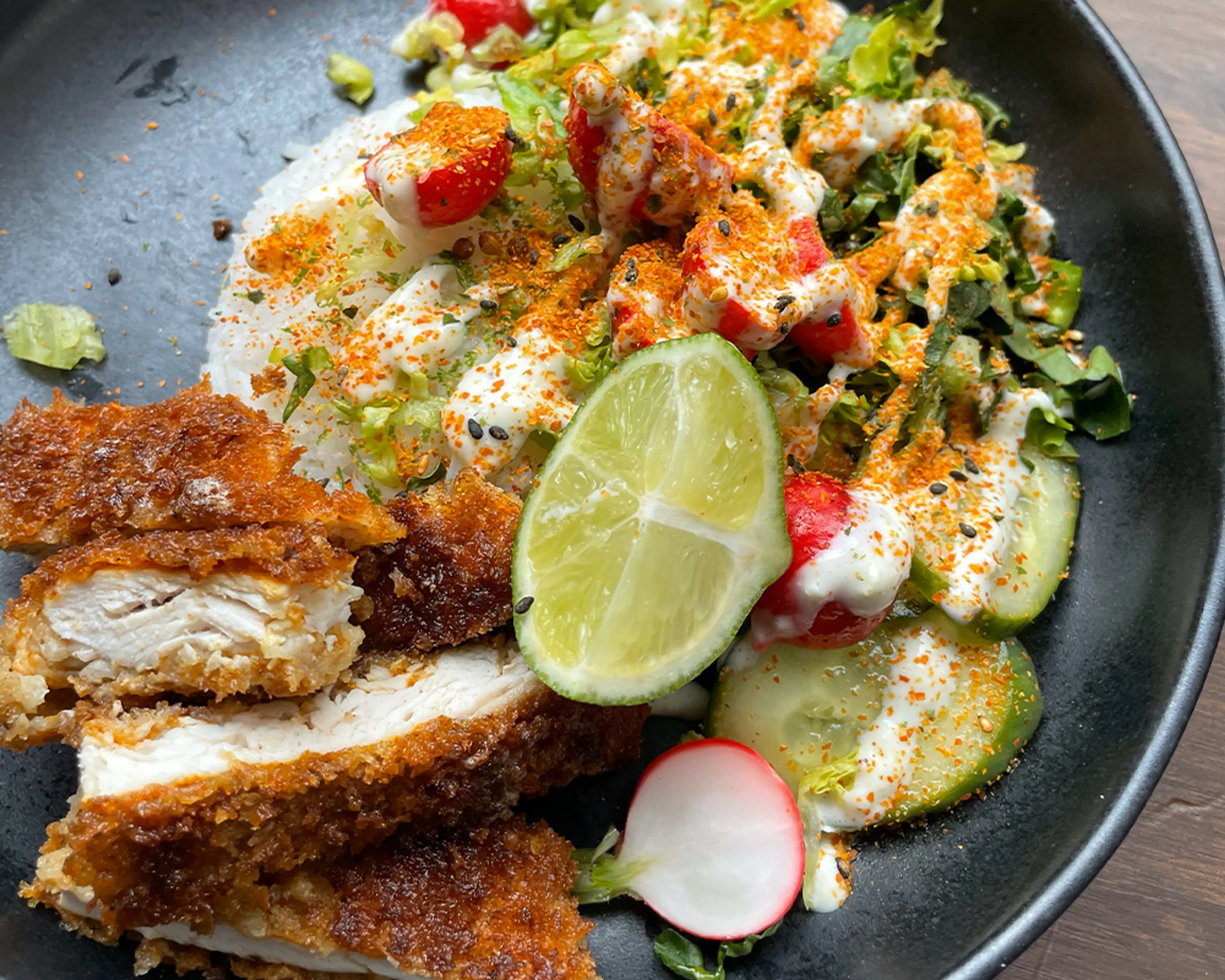 En tallrik med krispig, panerad kycklingschnitzel serverad med färsk sallad, skivade rädisor, gurka och en limeklyfta, toppad med en krämig sås och kryddor.