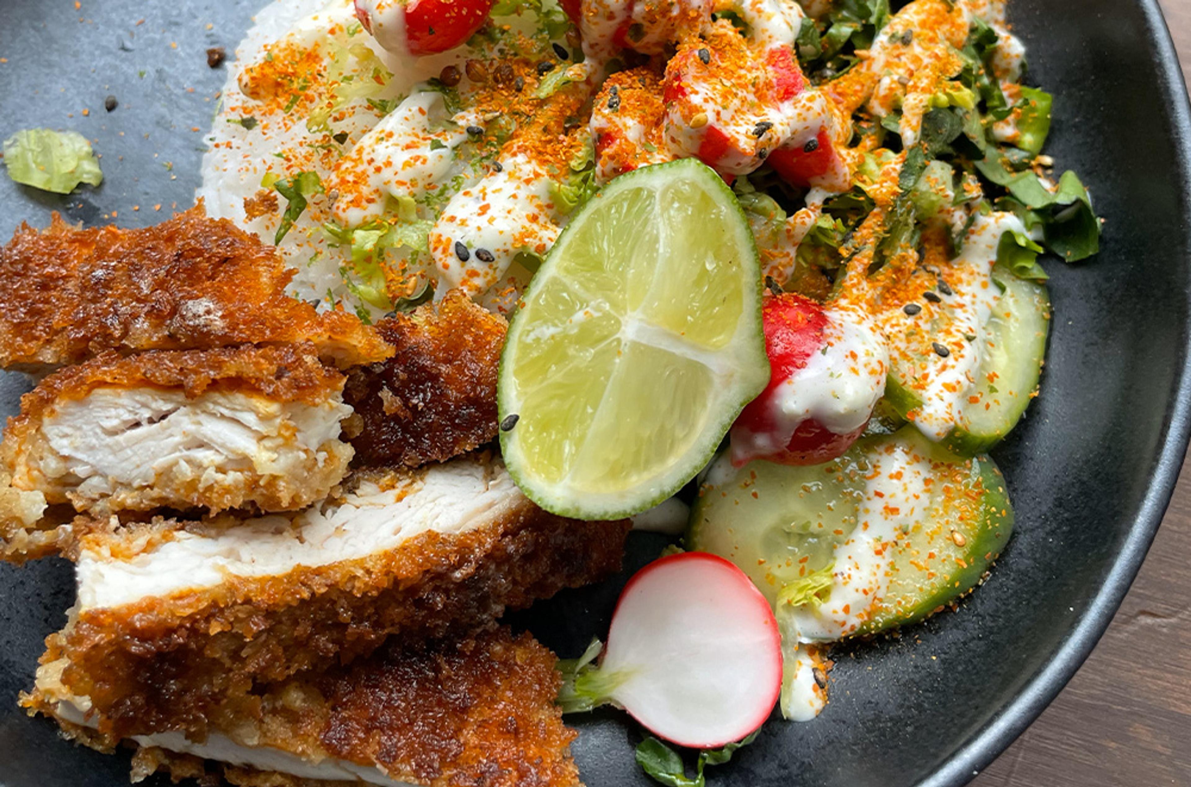 En tallrik med krispig, panerad kycklingschnitzel serverad med färsk sallad, skivade rädisor, gurka och en limeklyfta, toppad med en krämig sås och kryddor.