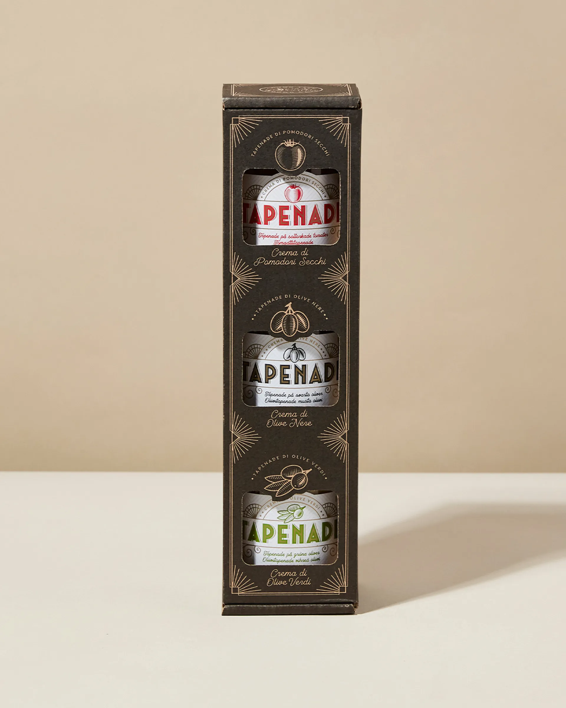 Tre små glasburkar med tapenade i en stilren, mörk förpackning, där smakerna röda tomater, svarta oliver och gröna oliver syns genom fönster på framsidan.