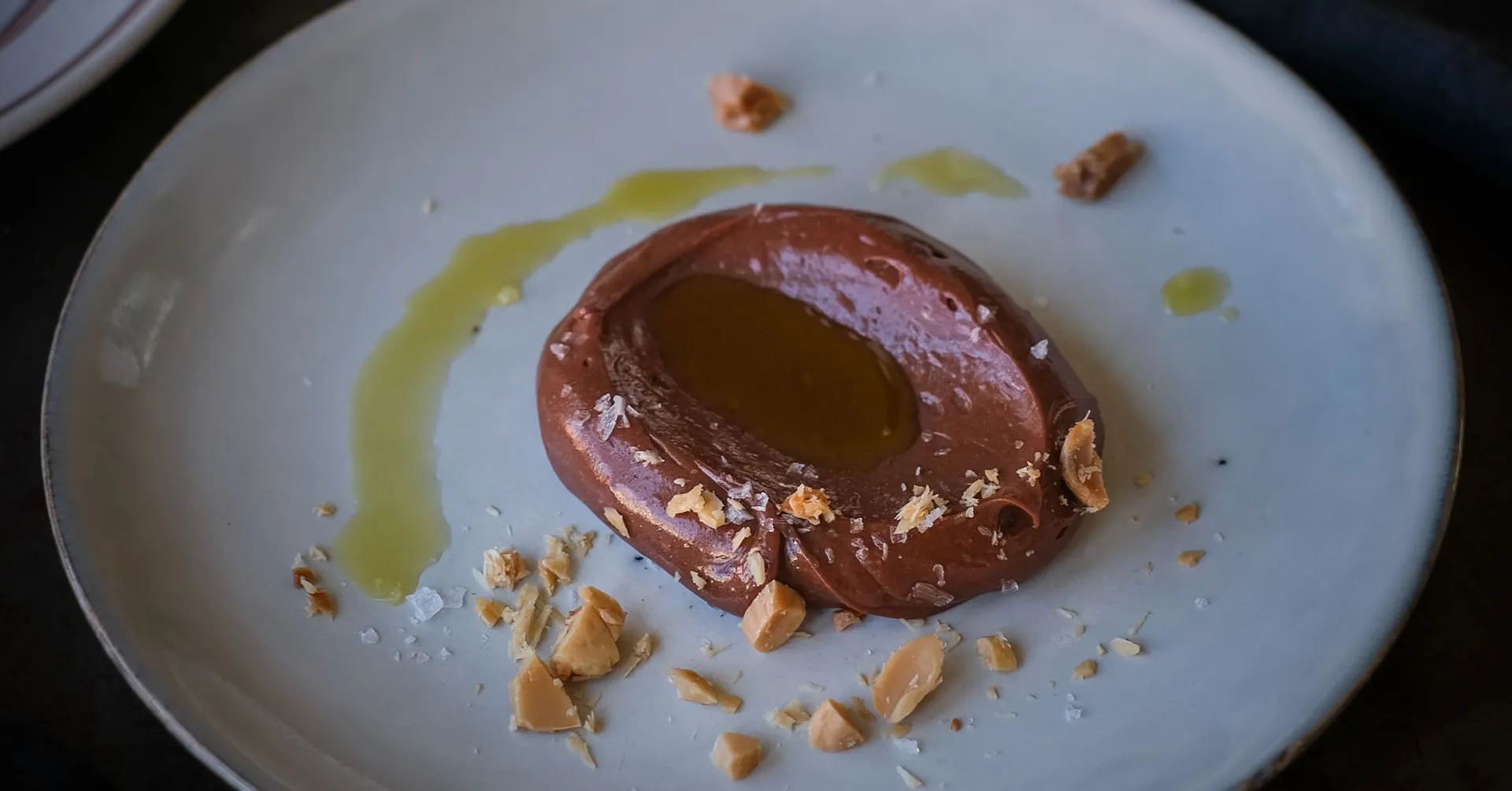 En klick len chokladkräm med en glansig olivolja i mitten, toppad med grovhackade nötbitar och ett stänk av flingsalt på en vit tallrik. I bakgrunden står en flaska extra virgin olivolja och en liten skål