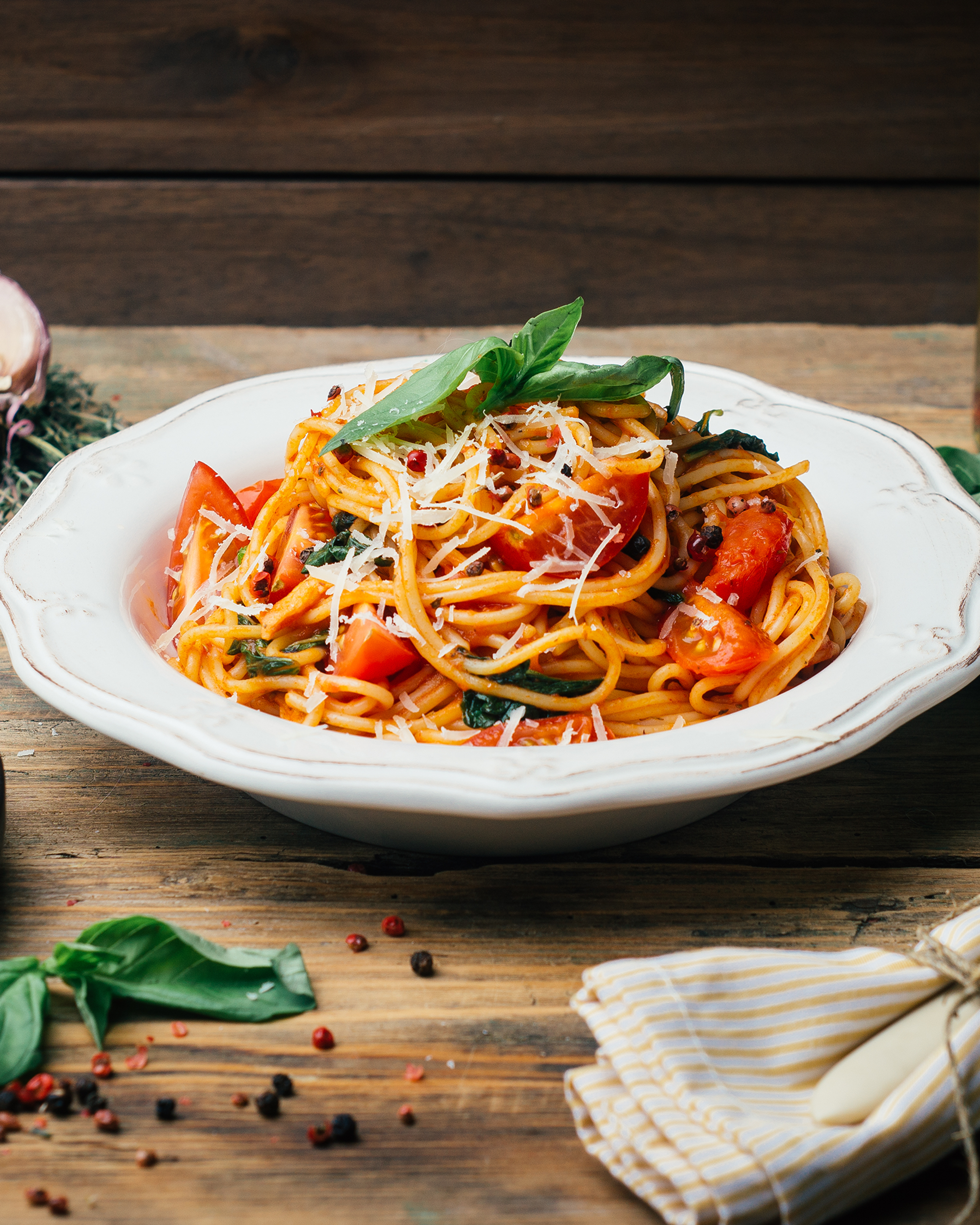 Spagetti med tomat, riven ost och färsk basilika i en vit djup tallrik på träbord. Kryddor och en vitgulrandig servett med bestick ligger bredvid.