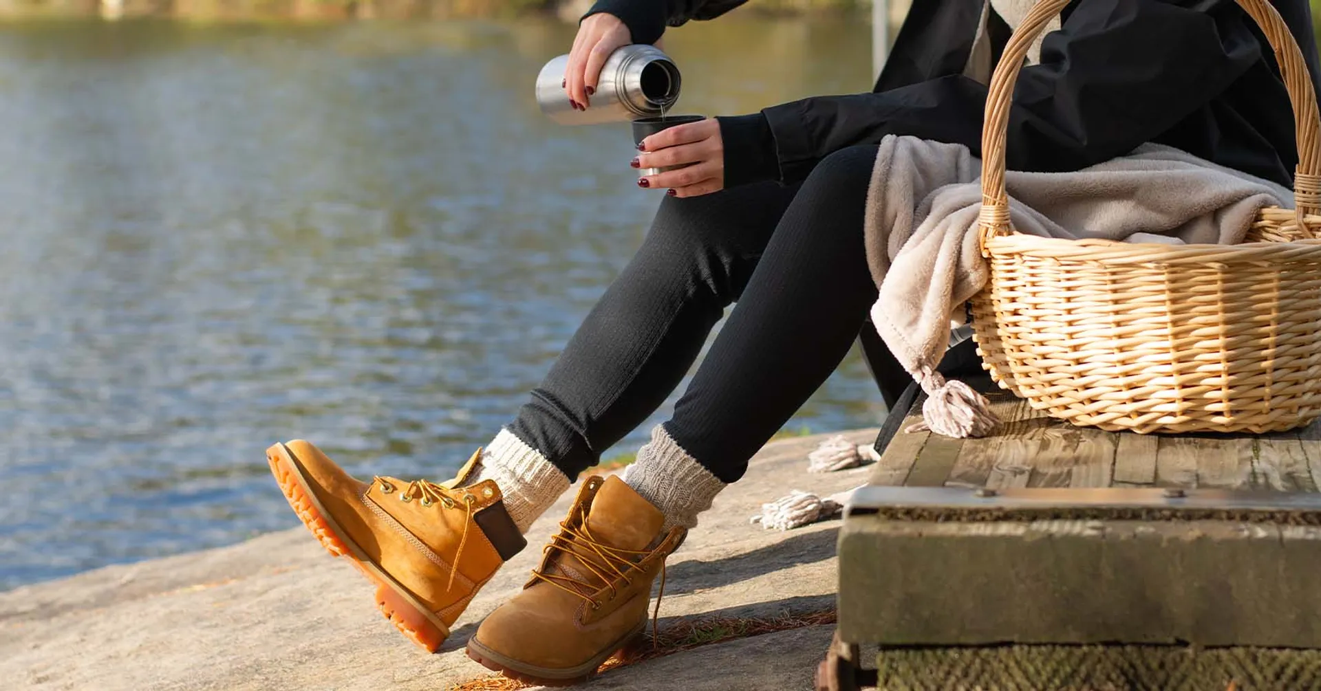 En person i svarta kläder och varma skor häller upp dryck från en termos vid en sjö. En flätad korg med filt står på träbryggan intill.