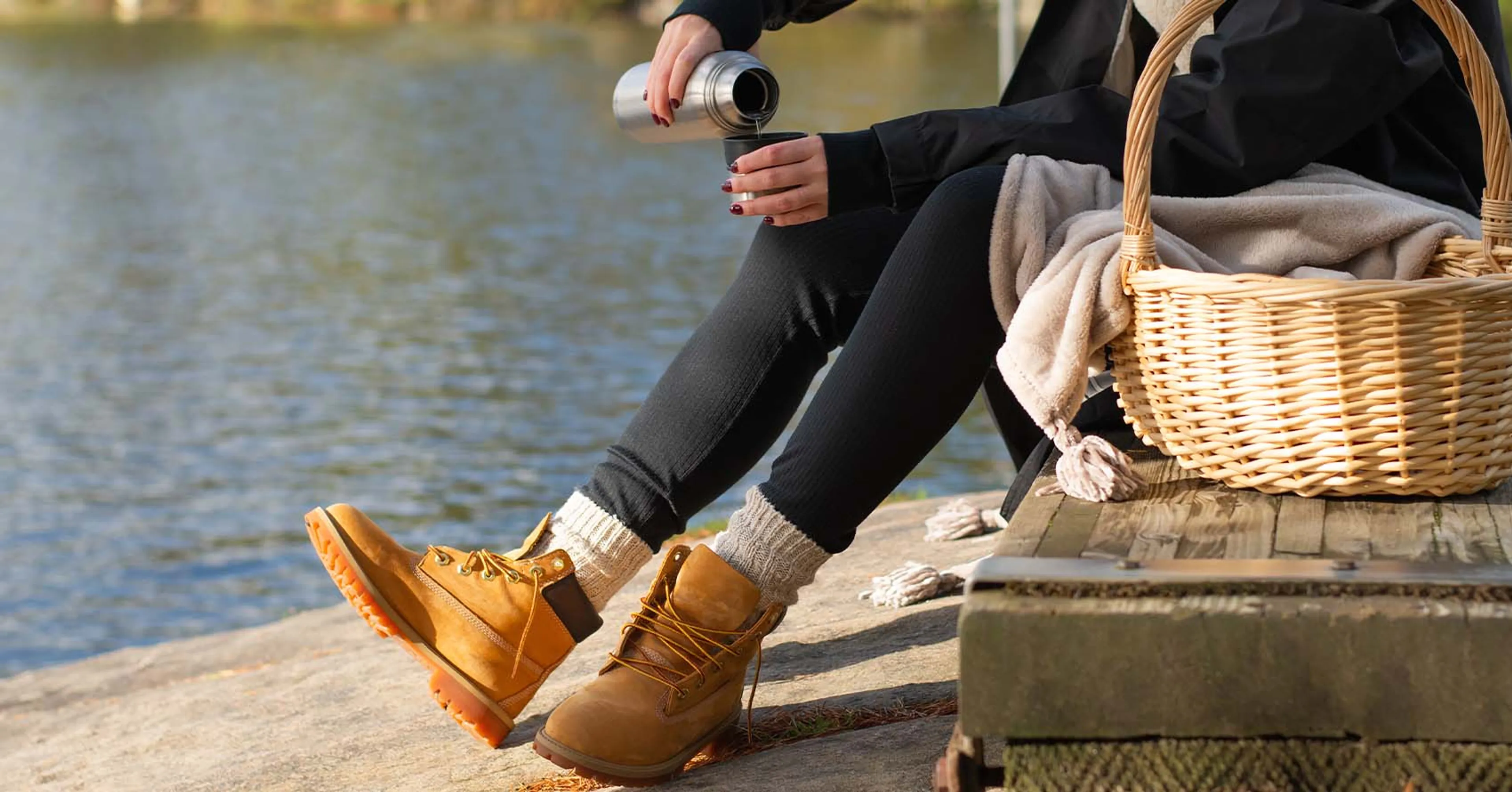 En person i svarta kläder och varma skor häller upp dryck från en termos vid en sjö. En flätad korg med filt står på träbryggan intill.