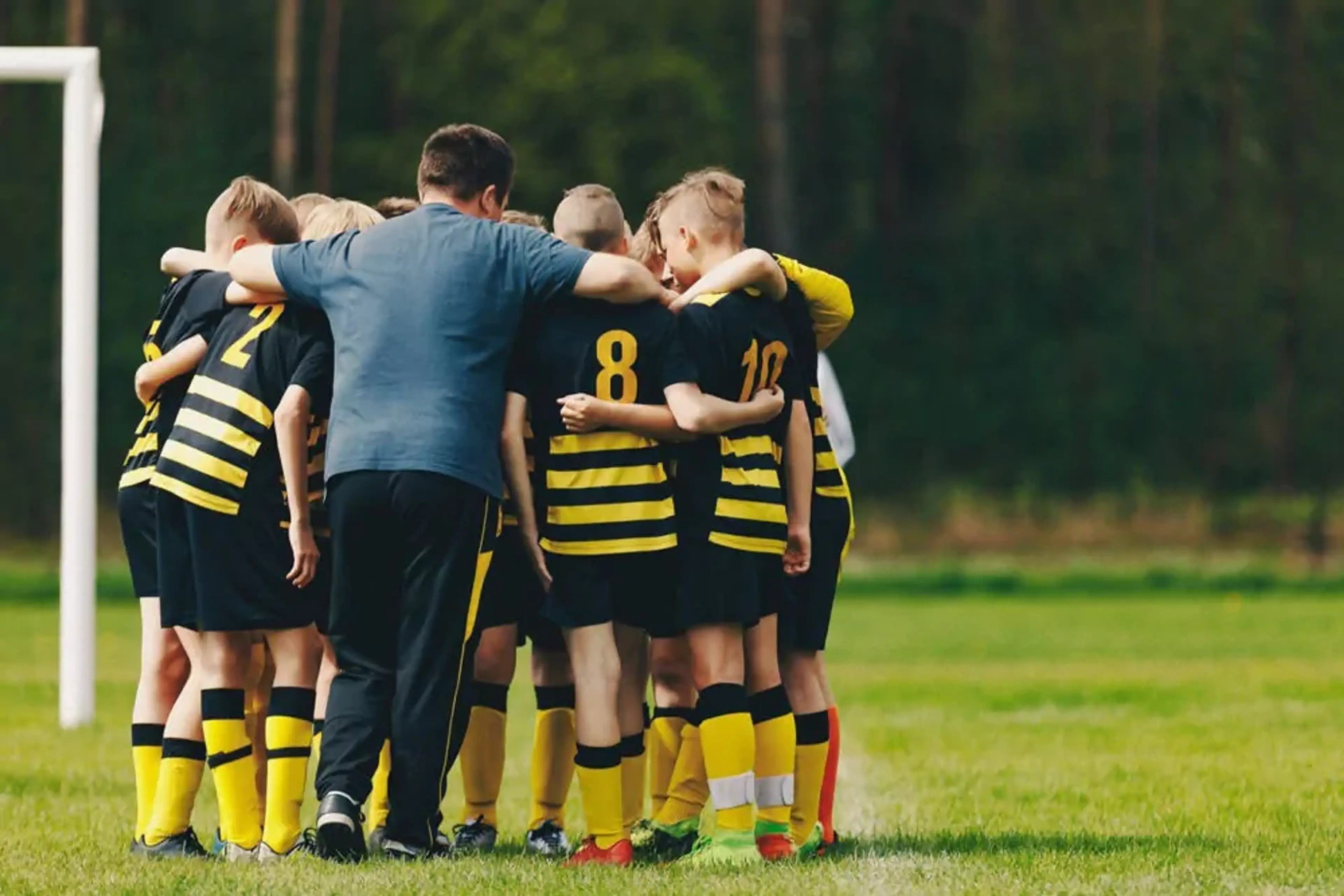 Ett ungdomslag i svart och gula fotbollströjor samlas i en cirkel tillsammans med en tränare på en grön fotbollsplan. Alla har armarna om varandra inför match.