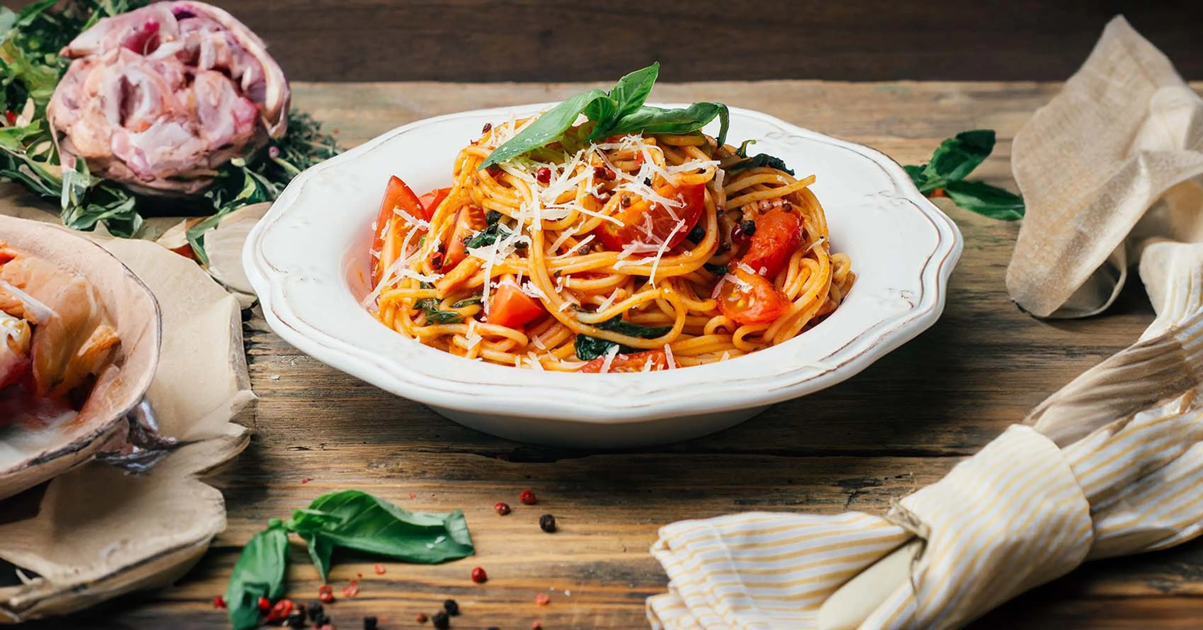 En tallrik med spaghetti blandad med tomater, basilika och riven ost. Pastan ligger upplagd på en vit tallrik mot en rustik träbakgrund.