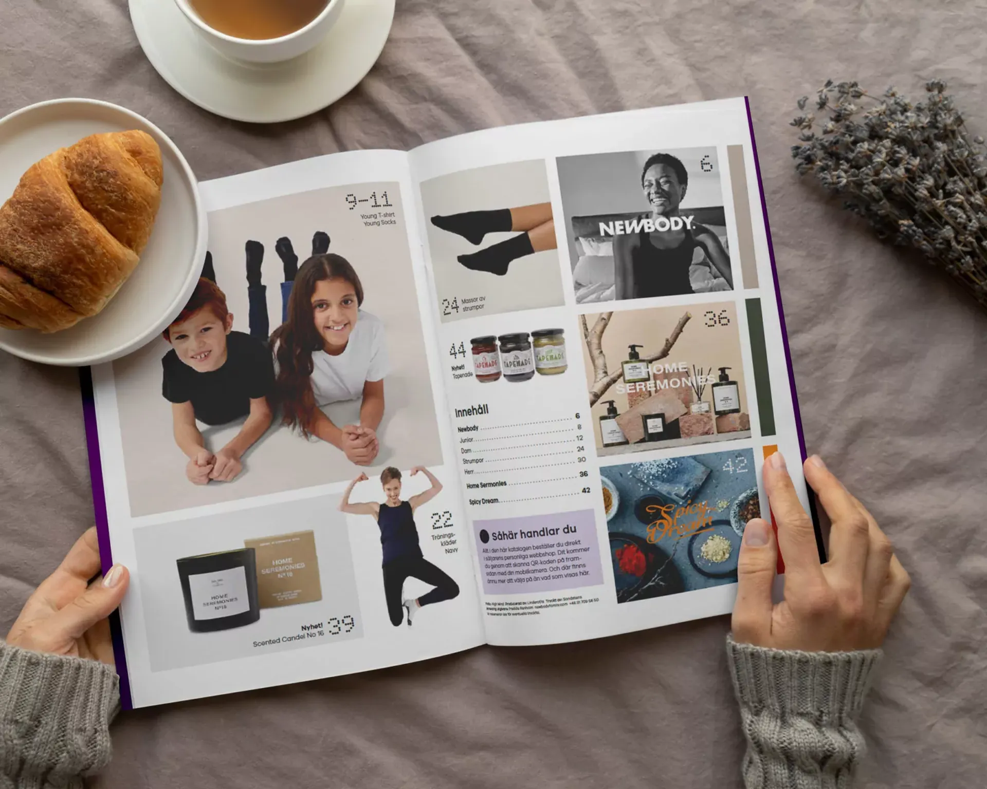En öppen katalog med bilder på barn i enkla kläder, träningskläder, doftljus, accessoarer och kryddor. Två händer håller i katalogen vid en kopp te och en croissant på ett mjukt tyg.