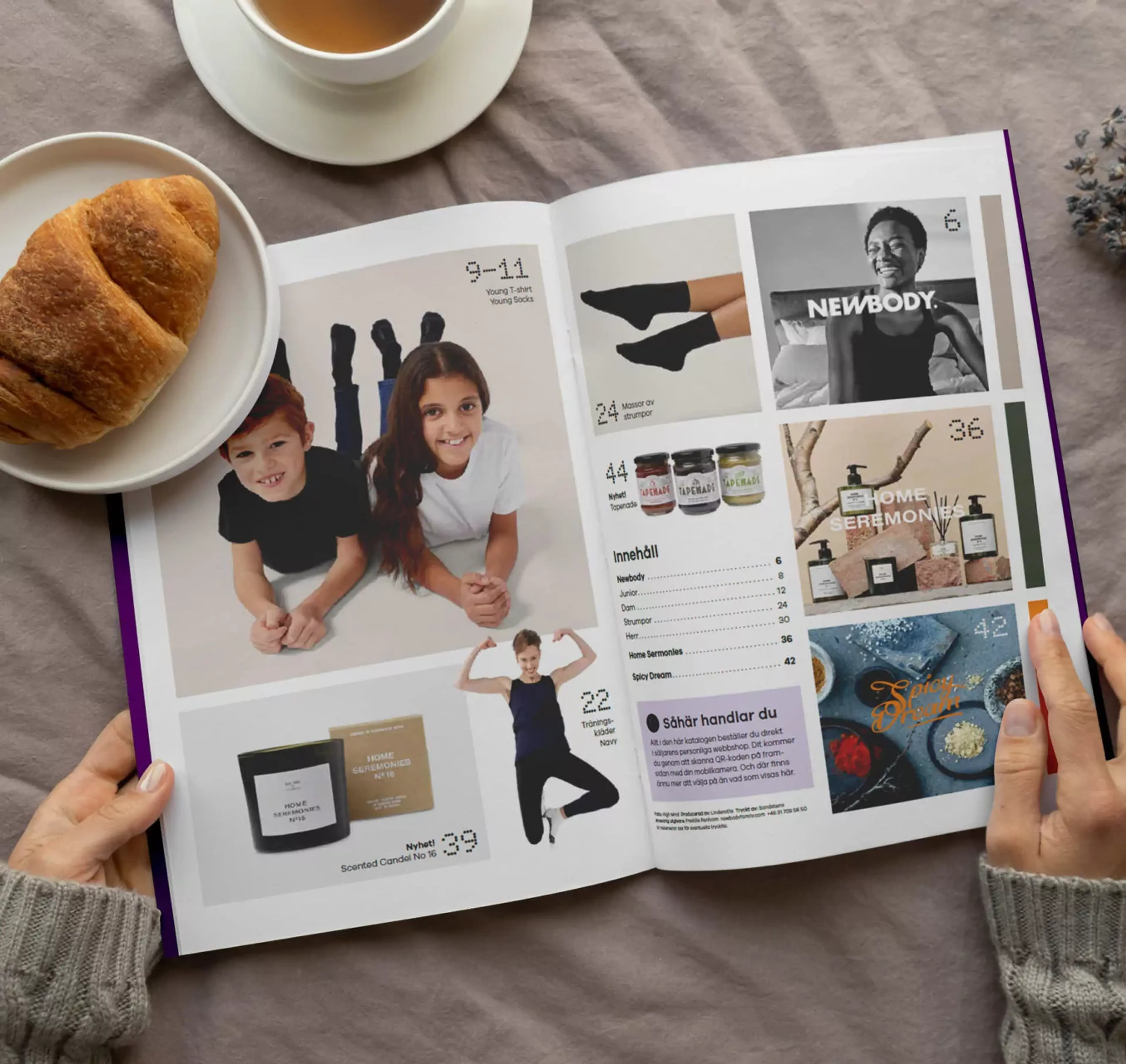 En öppen katalog med bilder på barn i enkla kläder, träningskläder, doftljus, accessoarer och kryddor. Två händer håller i katalogen vid en kopp te och en croissant på ett mjukt tyg.