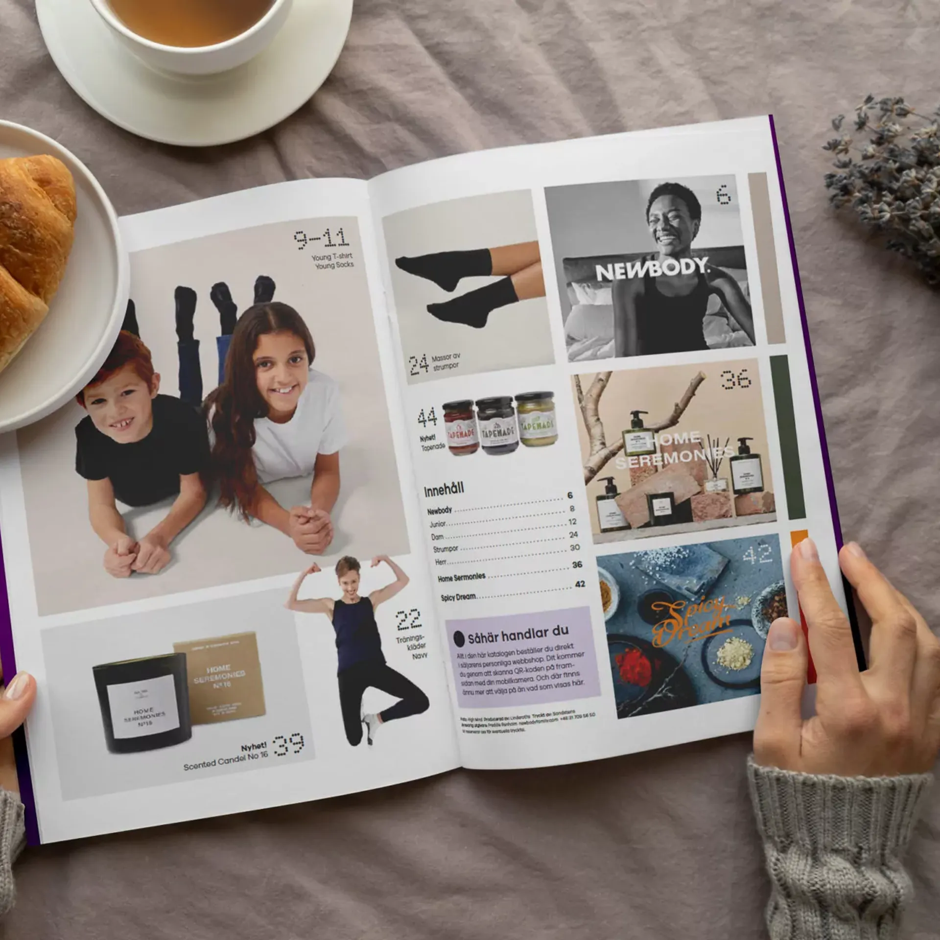 En öppen katalog med bilder på barn i enkla kläder, träningskläder, doftljus, accessoarer och kryddor. Två händer håller i katalogen vid en kopp te och en croissant på ett mjukt tyg.