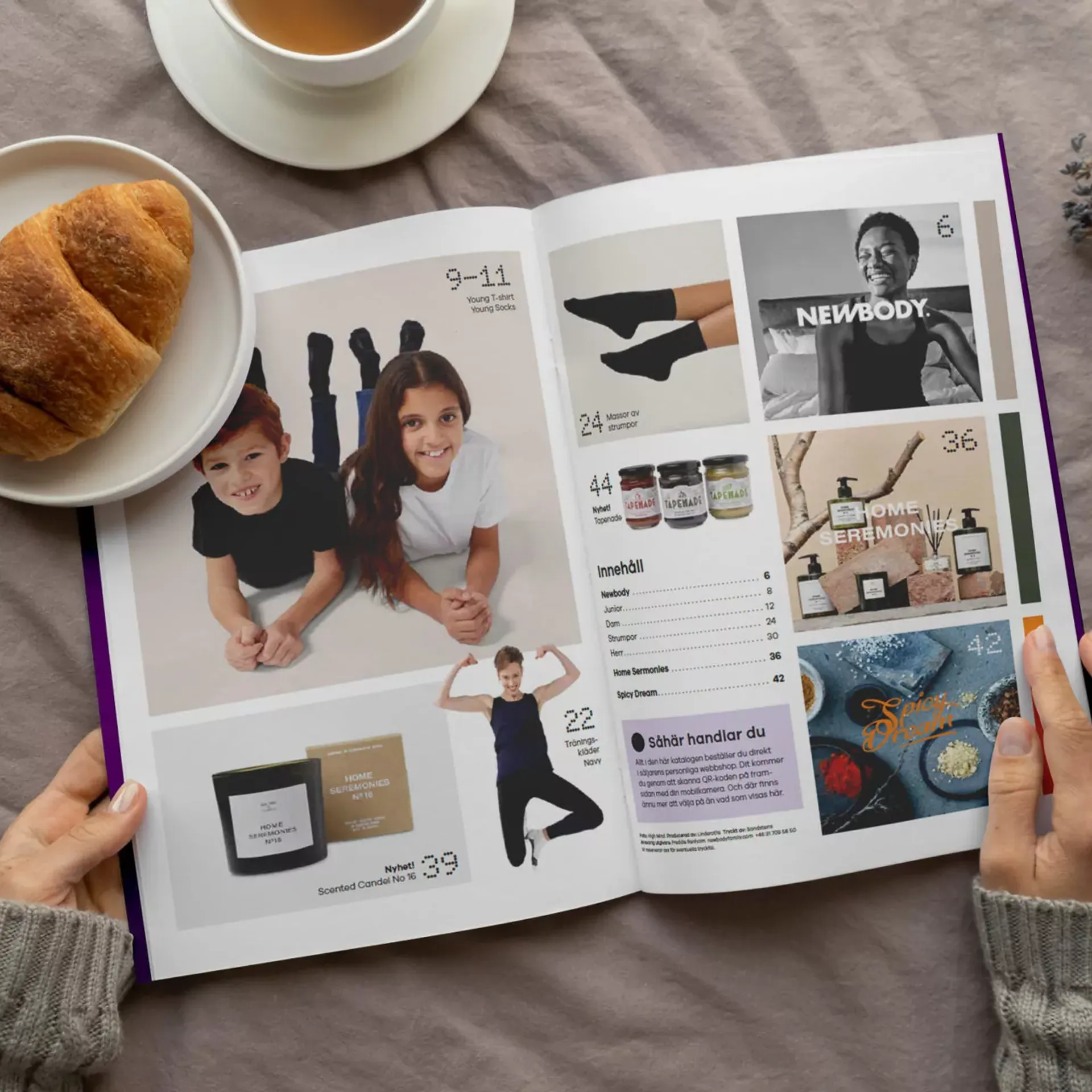En öppen katalog med bilder på barn i enkla kläder, träningskläder, doftljus, accessoarer och kryddor. Två händer håller i katalogen vid en kopp te och en croissant på ett mjukt tyg.