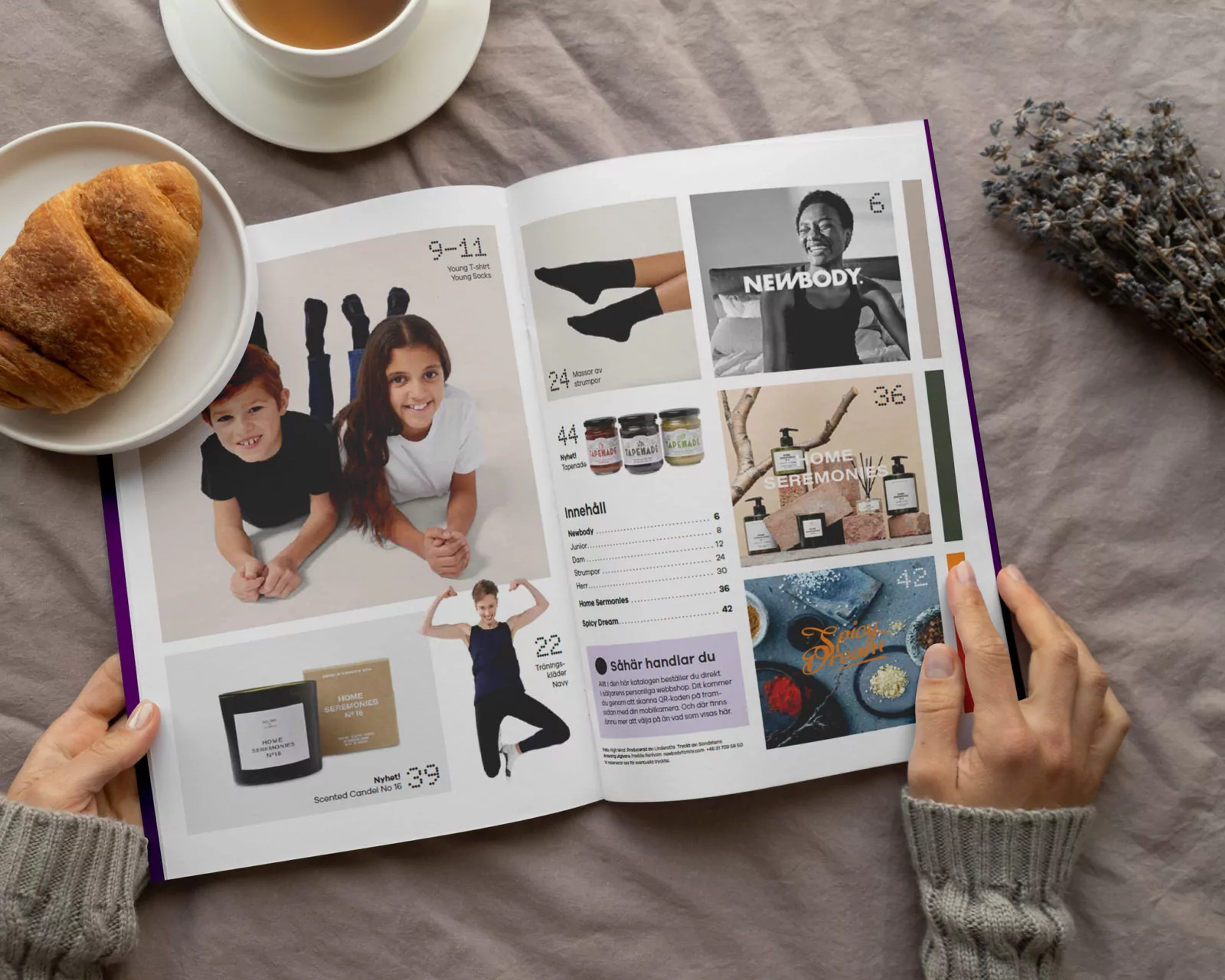 En öppen katalog med bilder på barn i enkla kläder, träningskläder, doftljus, accessoarer och kryddor. Två händer håller i katalogen vid en kopp te och en croissant på ett mjukt tyg.