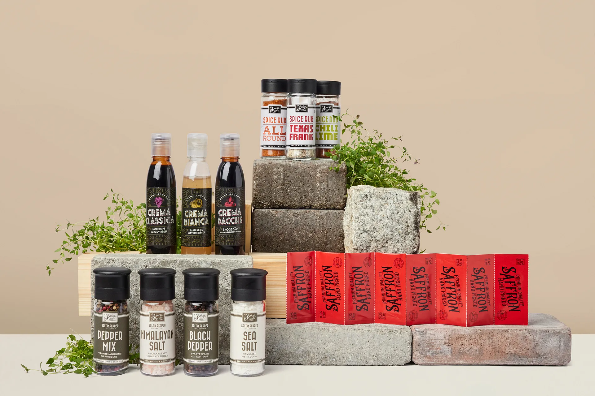 Olika kryddor och såser i flaskor och burkar, uppställda på stenblock med några gröna örter som dekoration. Sortimentet innehåller bland annat peppar, salt, saffran och smaksatta kryddblandningar.