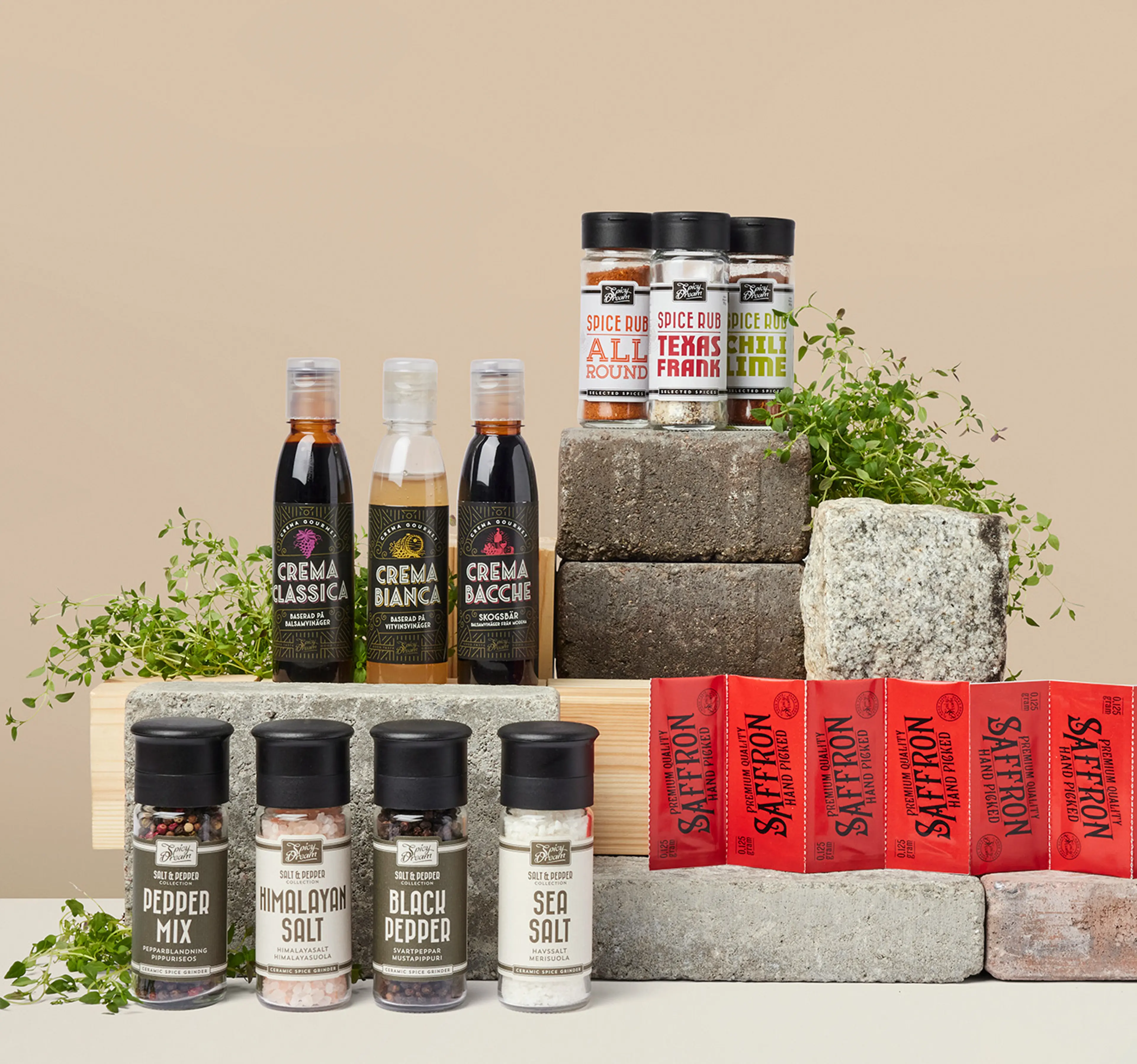 Olika kryddor och såser i flaskor och burkar, uppställda på stenblock med några gröna örter som dekoration. Sortimentet innehåller bland annat peppar, salt, saffran och smaksatta kryddblandningar.