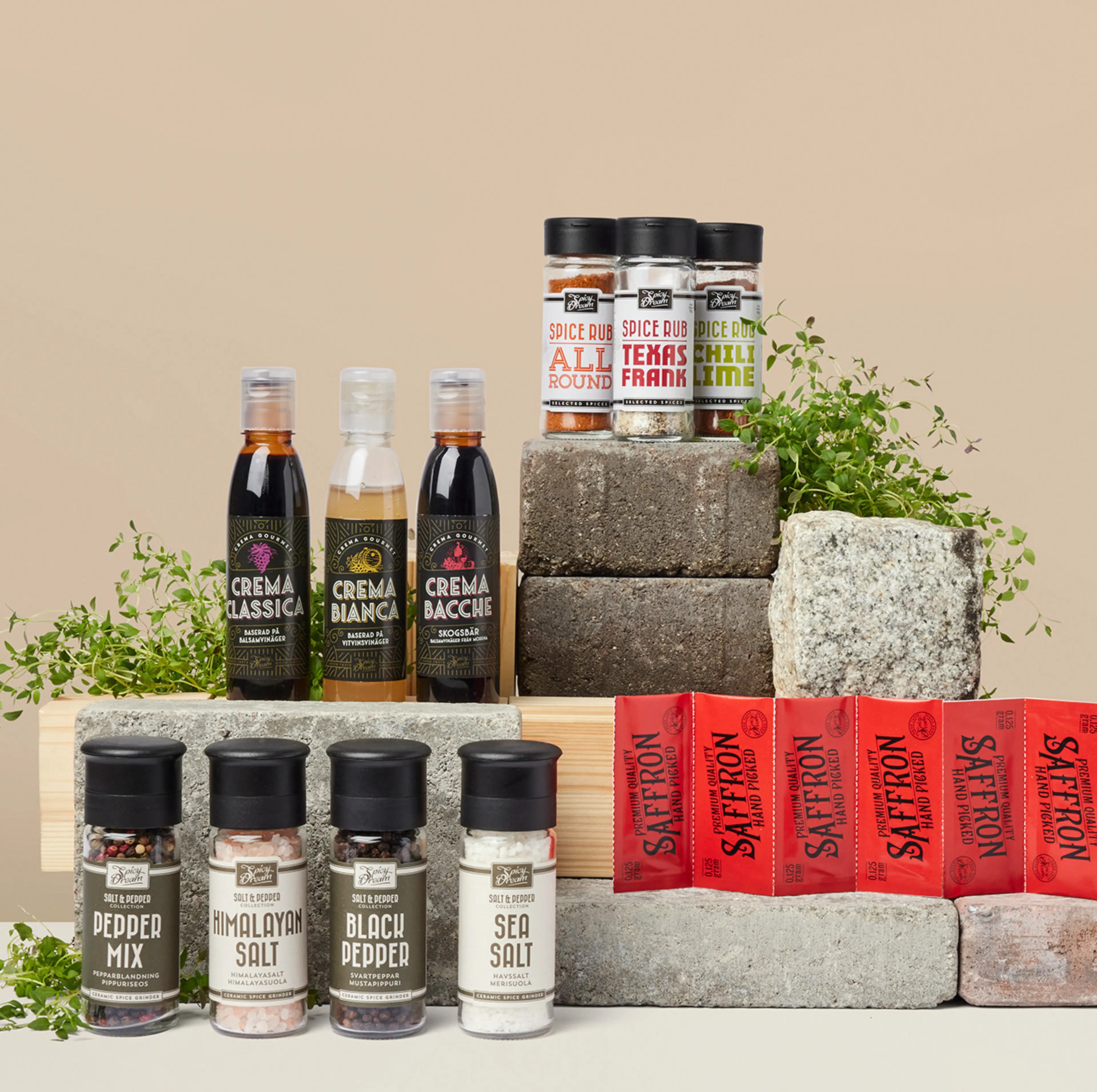 Olika kryddor och såser i flaskor och burkar, uppställda på stenblock med några gröna örter som dekoration. Sortimentet innehåller bland annat peppar, salt, saffran och smaksatta kryddblandningar.