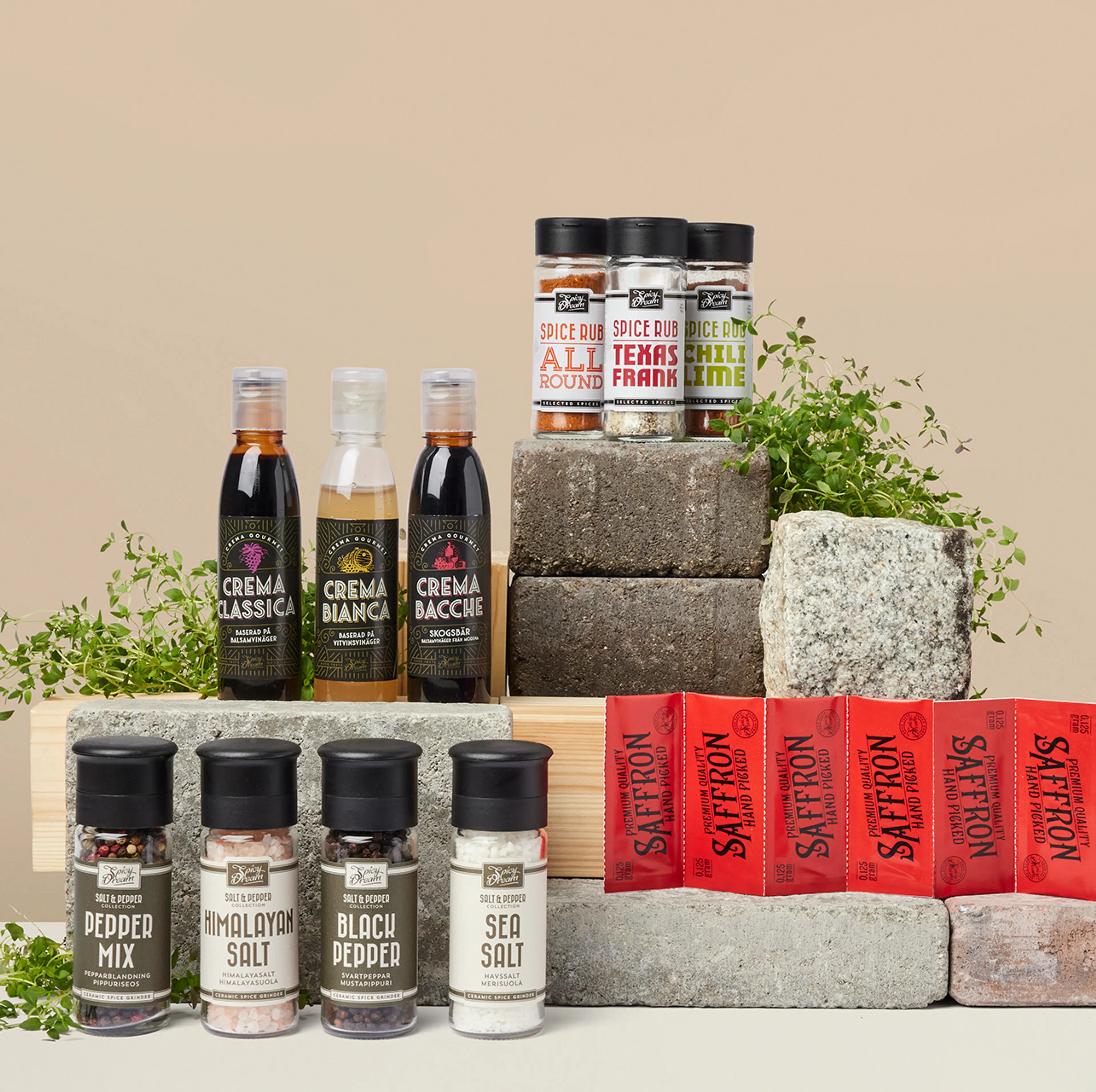 Olika kryddor och såser i flaskor och burkar, uppställda på stenblock med några gröna örter som dekoration. Sortimentet innehåller bland annat peppar, salt, saffran och smaksatta kryddblandningar.