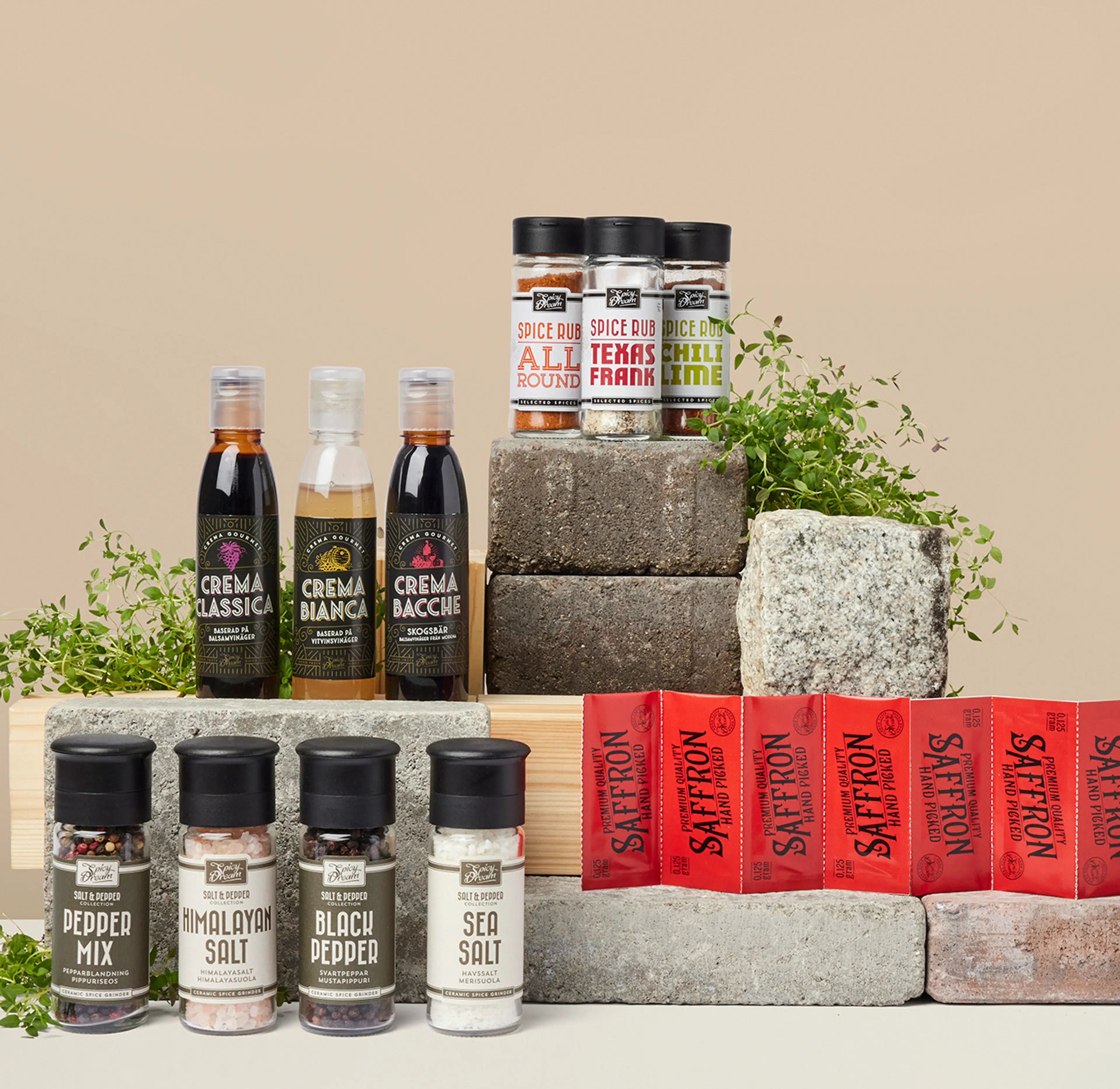 Olika kryddor och såser i flaskor och burkar, uppställda på stenblock med några gröna örter som dekoration. Sortimentet innehåller bland annat peppar, salt, saffran och smaksatta kryddblandningar.