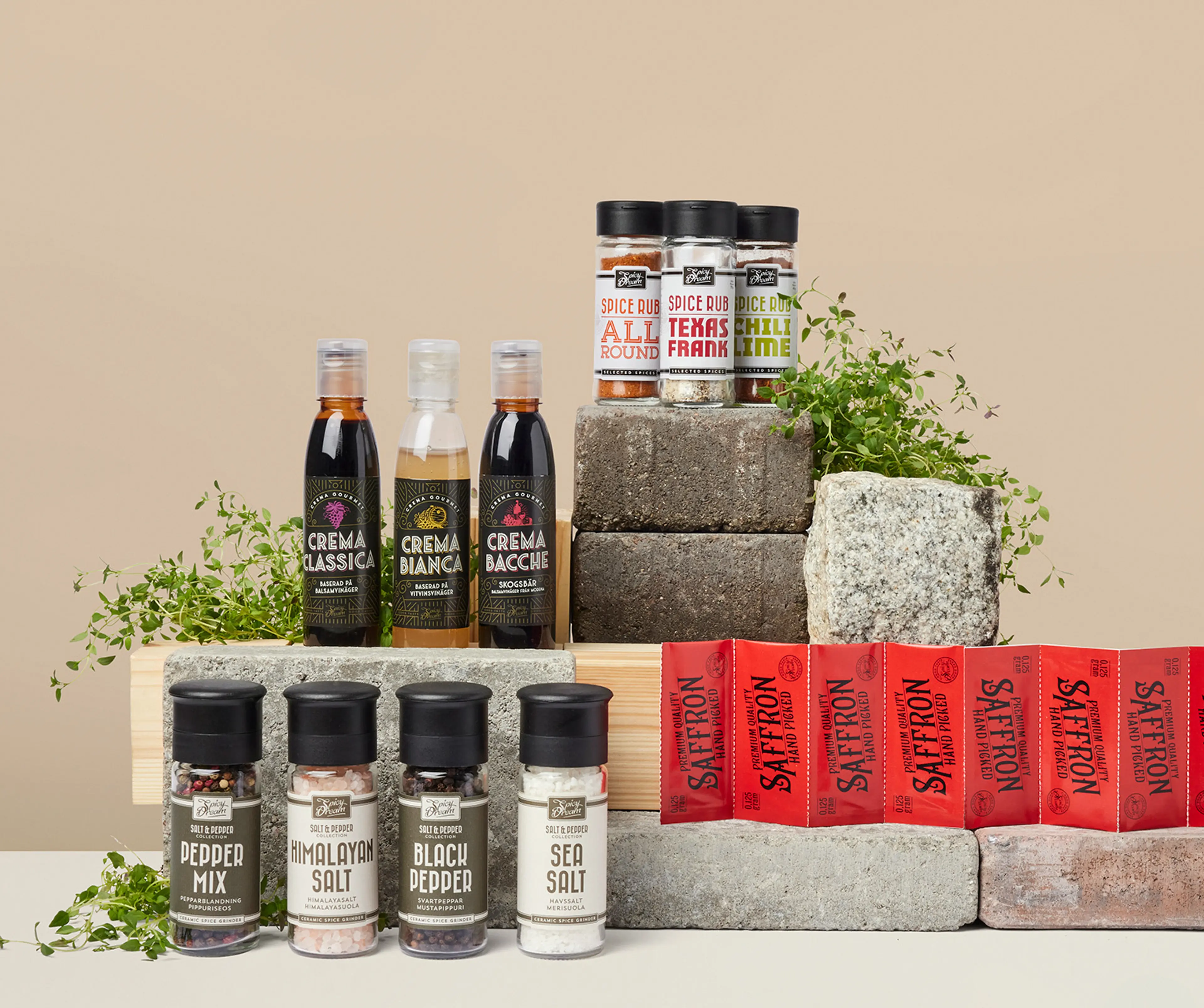 Olika kryddor och såser i flaskor och burkar, uppställda på stenblock med några gröna örter som dekoration. Sortimentet innehåller bland annat peppar, salt, saffran och smaksatta kryddblandningar.