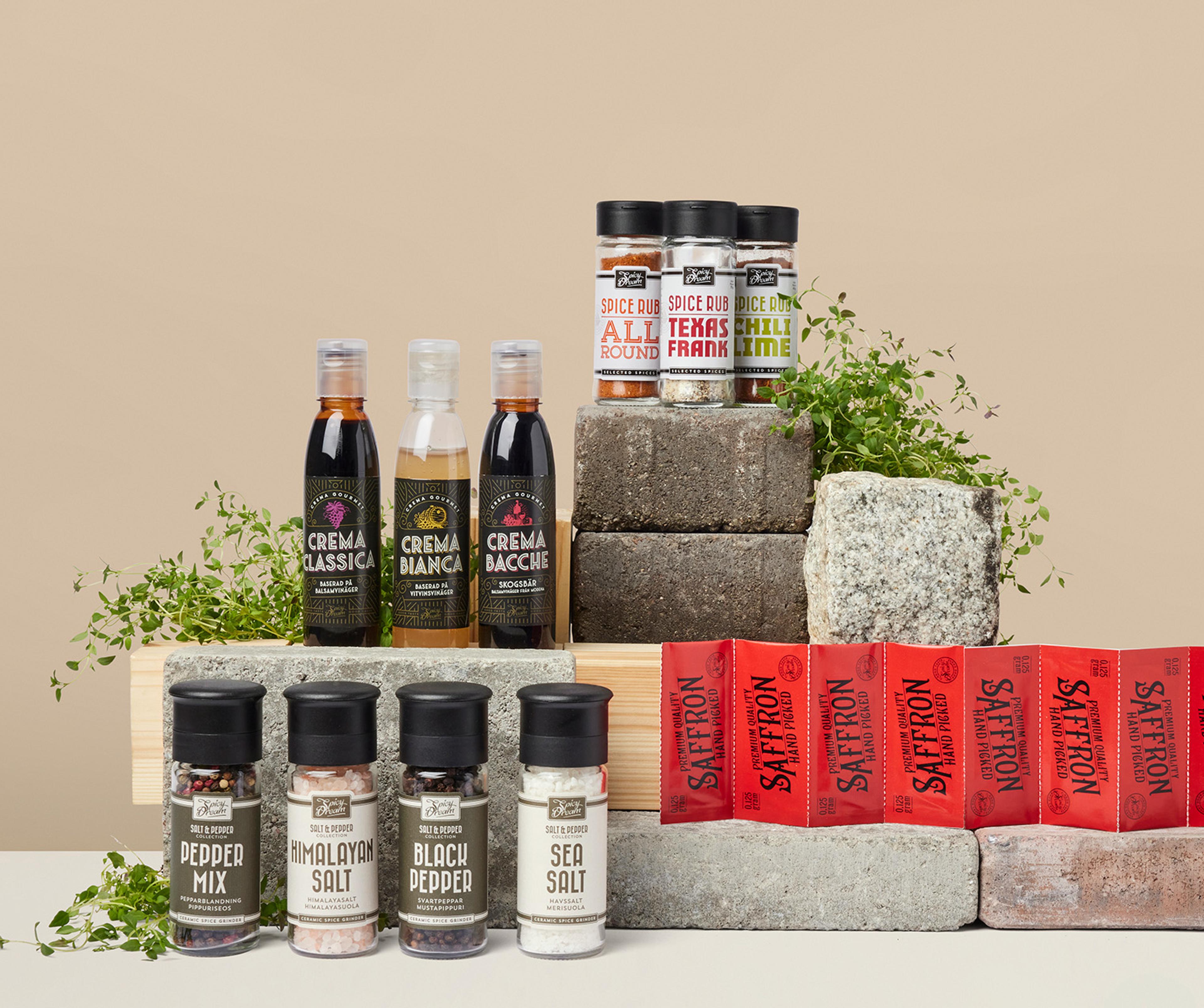 Olika kryddor och såser i flaskor och burkar, uppställda på stenblock med några gröna örter som dekoration. Sortimentet innehåller bland annat peppar, salt, saffran och smaksatta kryddblandningar.