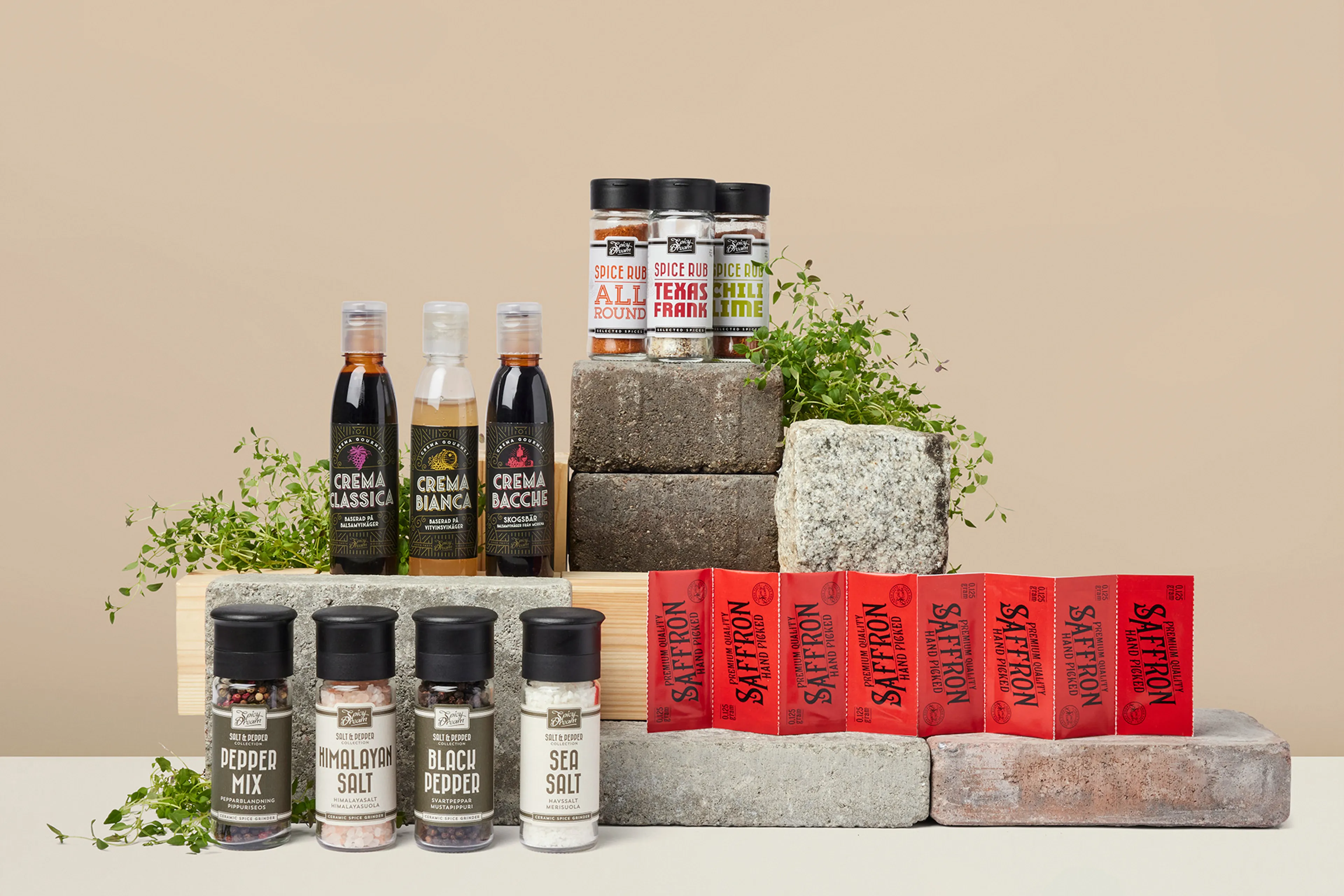 Olika kryddor och såser i flaskor och burkar, uppställda på stenblock med några gröna örter som dekoration. Sortimentet innehåller bland annat peppar, salt, saffran och smaksatta kryddblandningar.