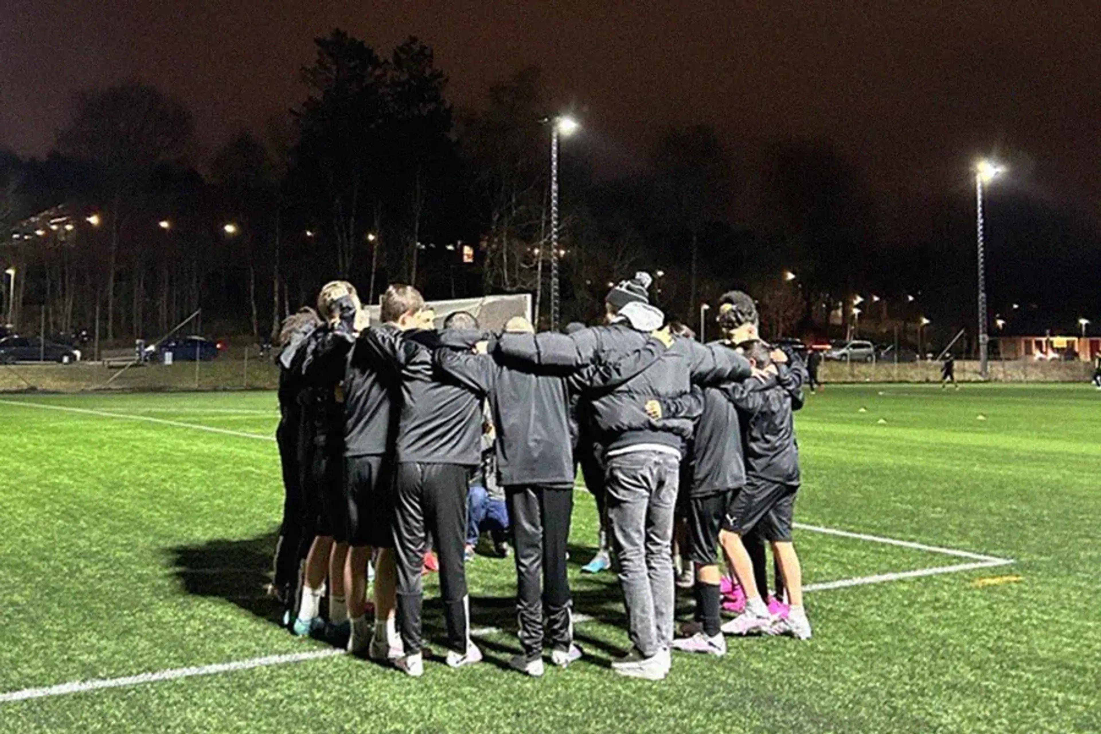 Ett fotbollslag står i en ring på en upplyst gräsplan under kvällen, med armarna runt varandra. Bakgrunden visar träd och några bilar i dunklet.