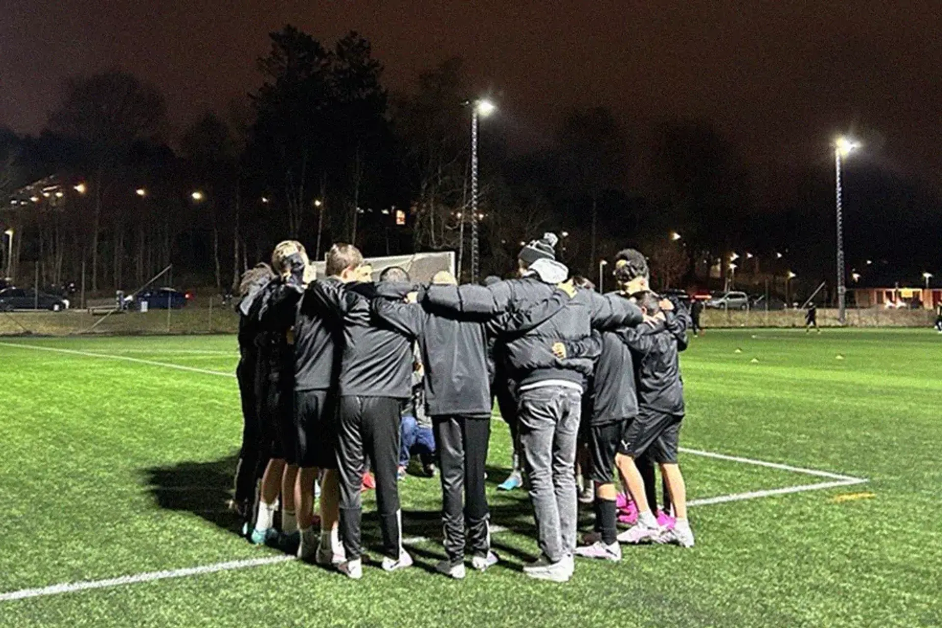 Ett fotbollslag står i en ring på en upplyst gräsplan under kvällen, med armarna runt varandra. Bakgrunden visar träd och några bilar i dunklet.