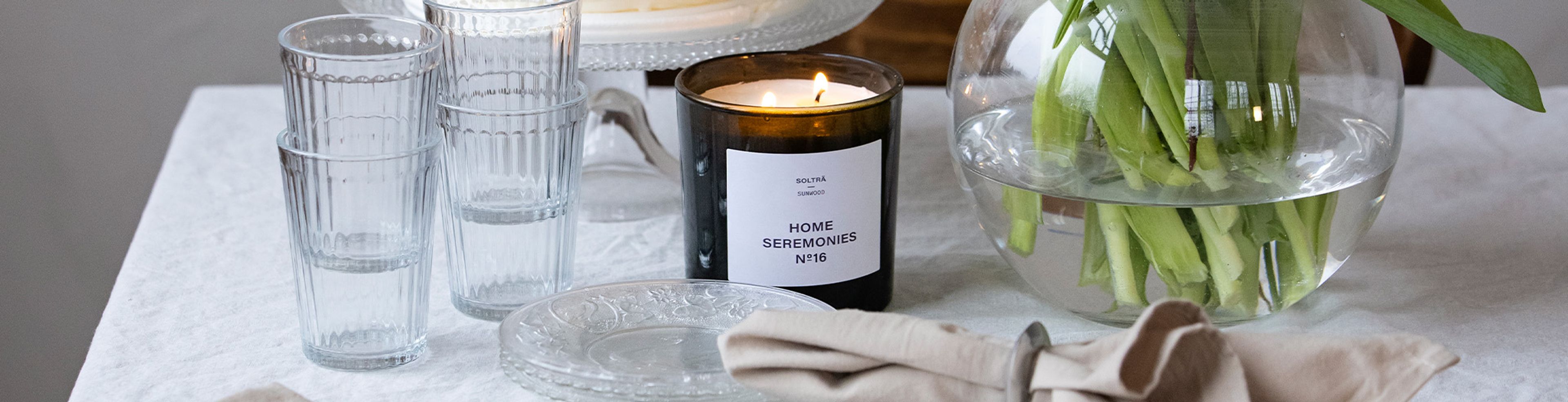 Ett svart doftljus märkt "Home Seremonies" brinner bredvid en rund glasvas med gröna stjälkar, omgivna av klara glas och en vacker glasskål på ett ljust dukat bord.
