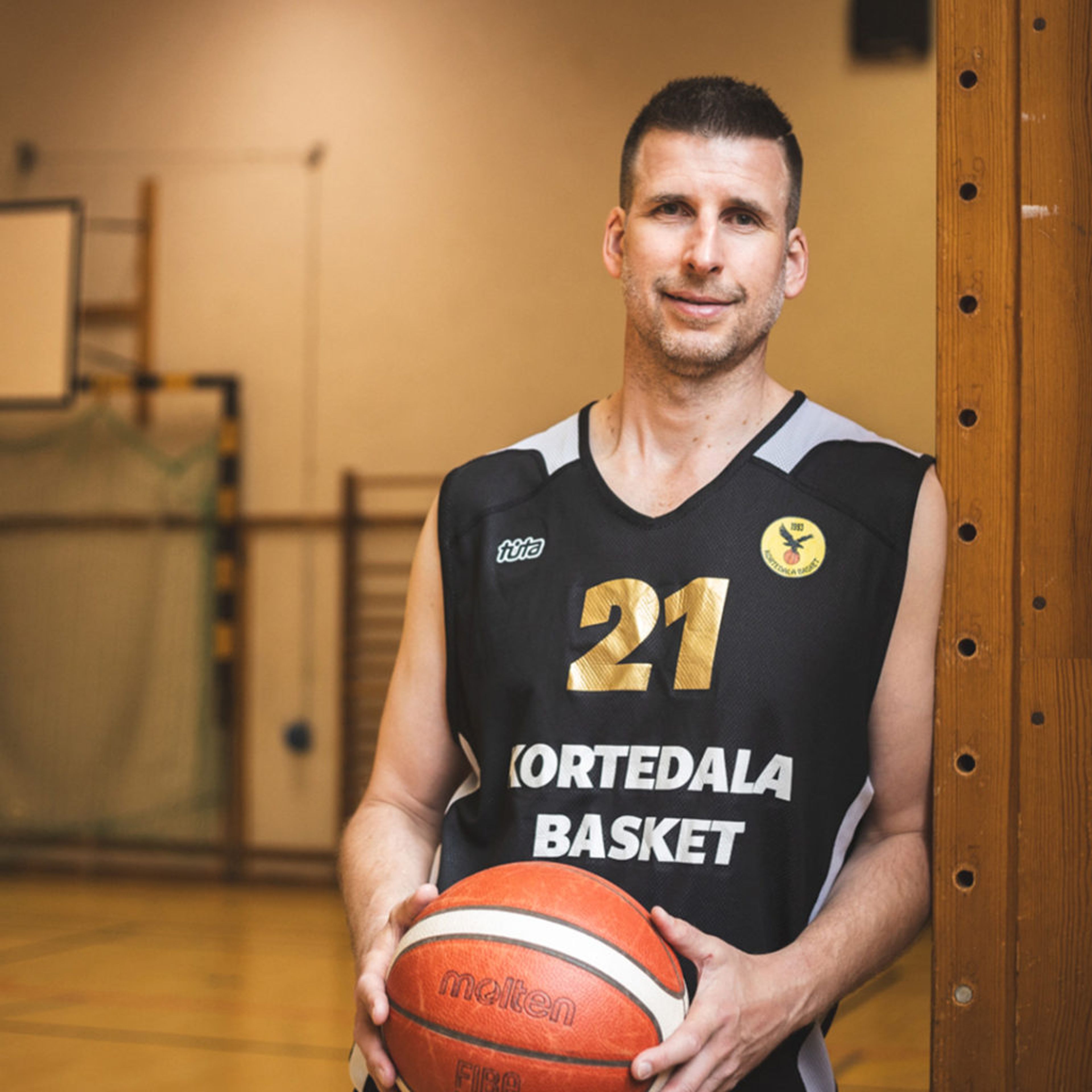 En man i svart baskettröja med nummer 21 och texten "Kortedala Basket" håller en basketboll i en sporthall. Bakgrunden visar målbur och basketkorg.