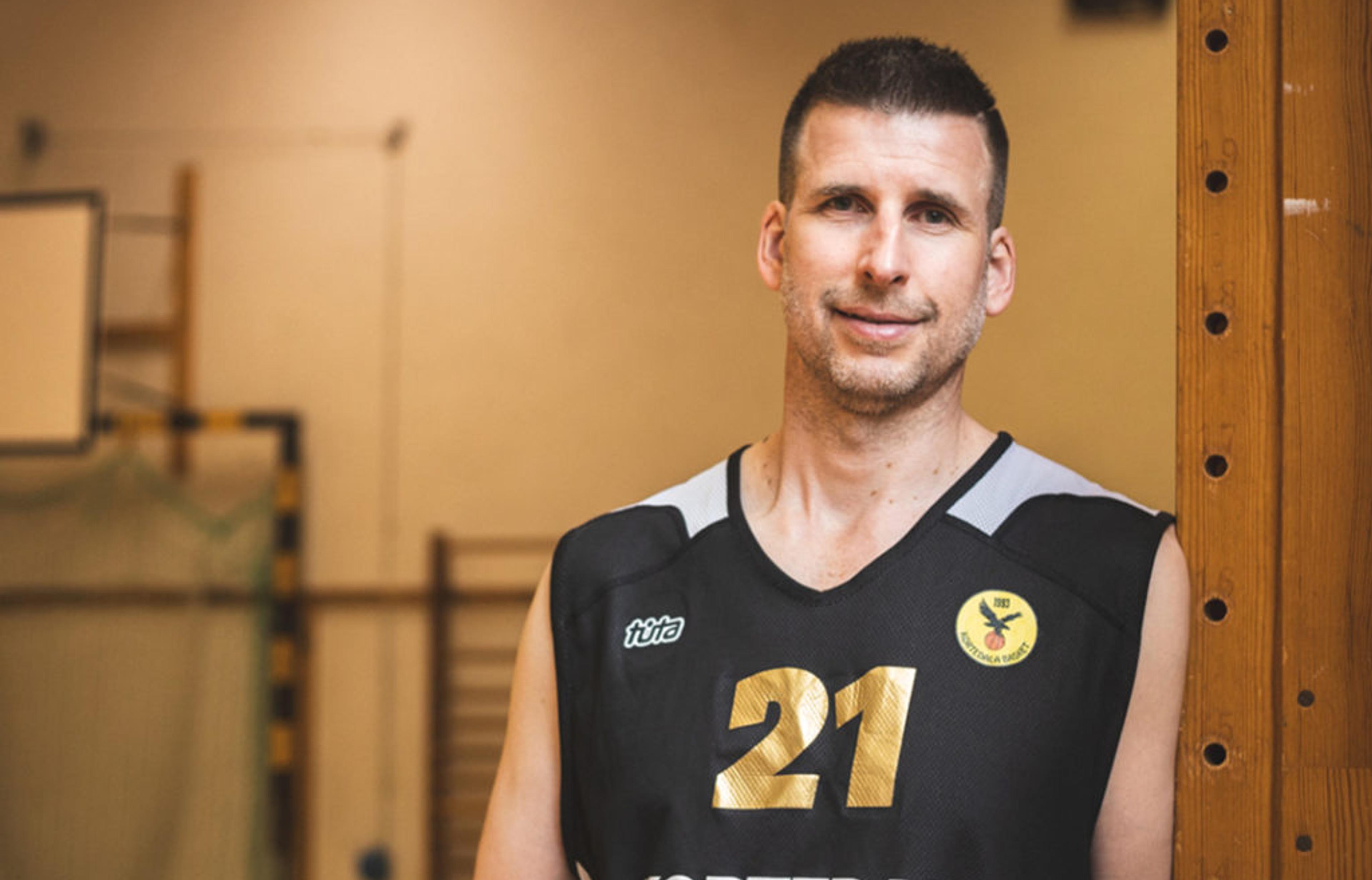 En man i svart baskettröja med nummer 21 och texten "Kortedala Basket" håller en basketboll i en sporthall. Bakgrunden visar målbur och basketkorg.