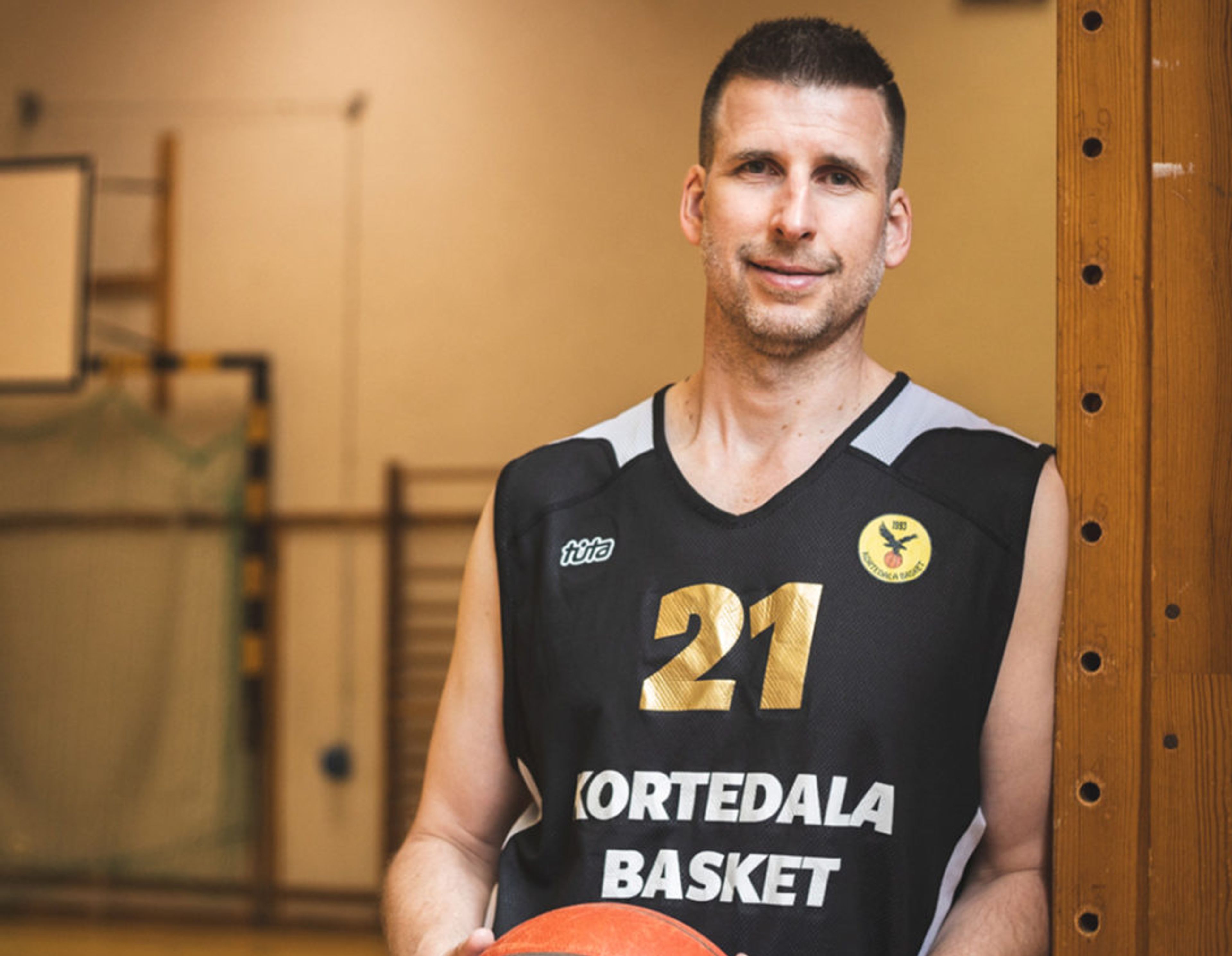 En man i svart baskettröja med nummer 21 och texten "Kortedala Basket" håller en basketboll i en sporthall. Bakgrunden visar målbur och basketkorg.