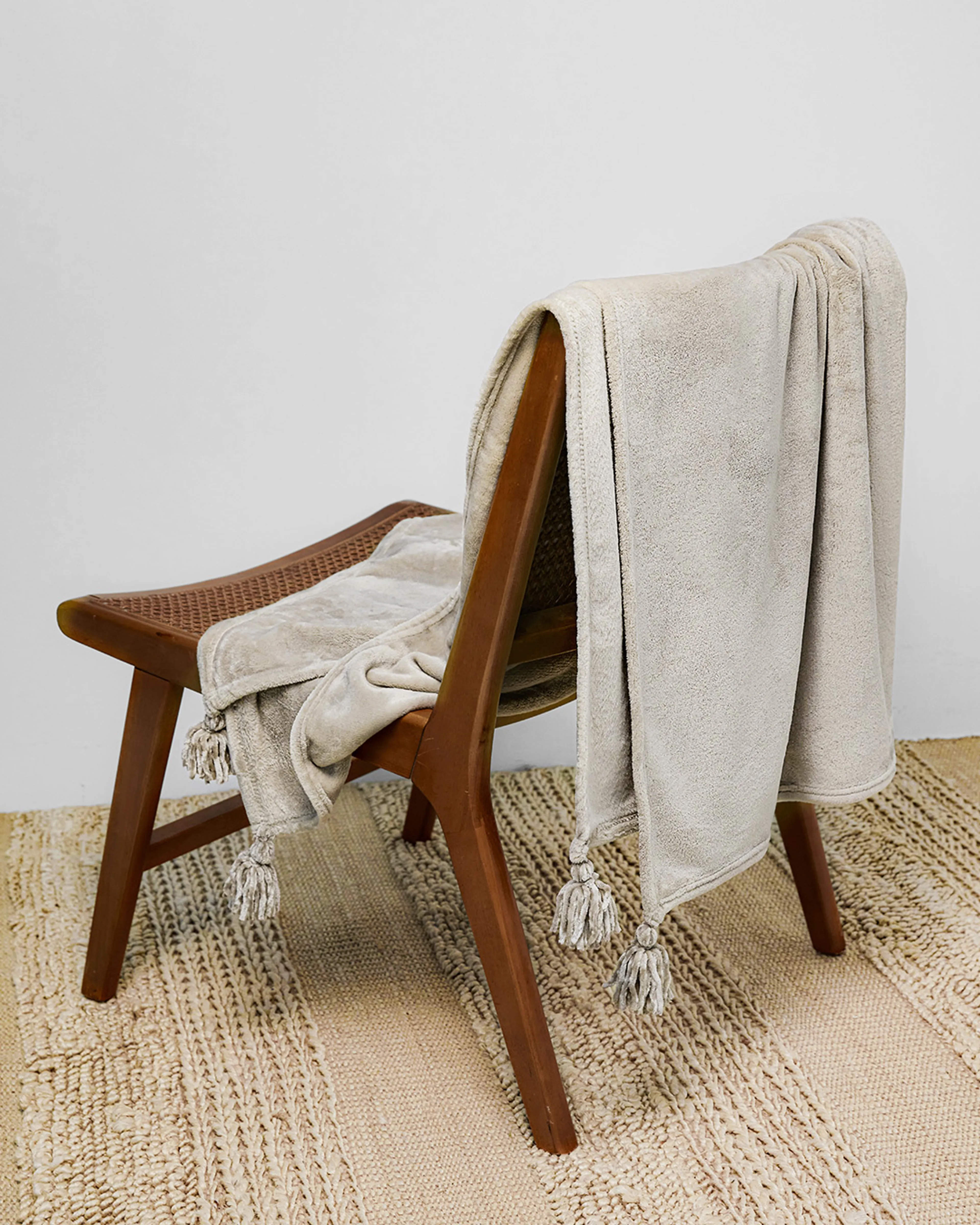 En mjuk, ljusgrå fleecefilt med dekorativa tofsar som hänger över ryggen på en enkel trästol. Filten ligger delvis utbredd över ett naturfärgat, grovt mattunderlag.