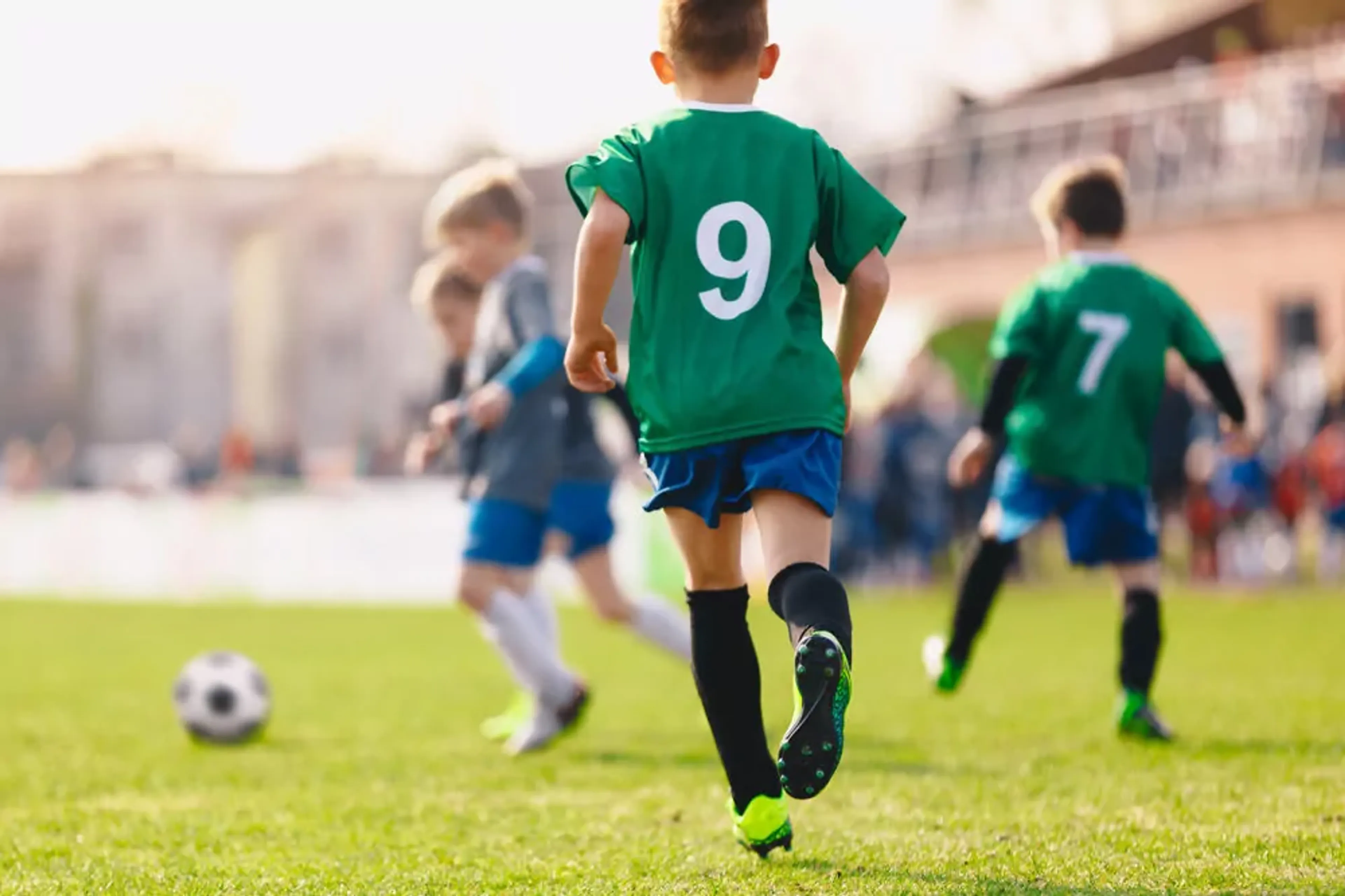 Barn i gröna tröjor och blå shorts spelar fotboll på en grön gräsmatta under en solig dag. Två av spelarna har nummer 9 respektive 7 på ryggen.