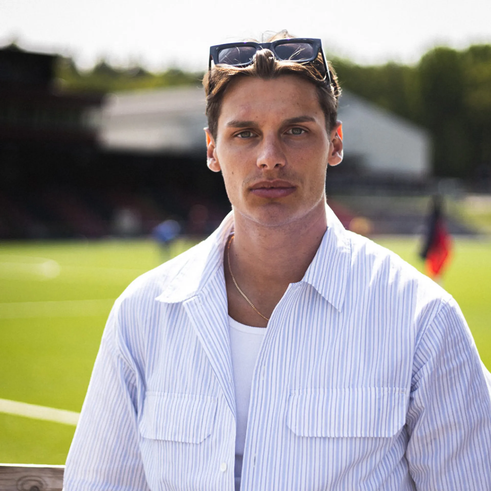 En ung man med solglasögon i håret bär en ljusblå, randig skjorta över en vit t-shirt. Bakgrunden visar en grön fotbollsplan i solljus.