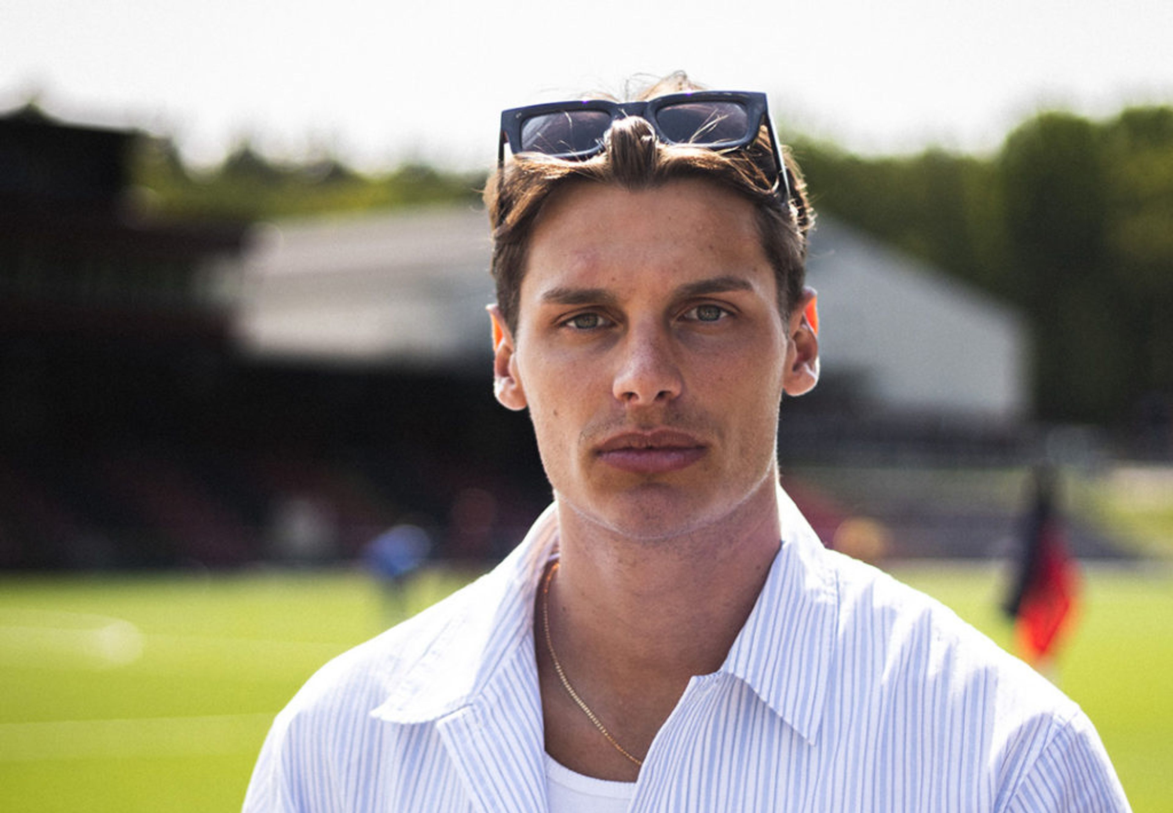 En ung man med solglasögon i håret bär en ljusblå, randig skjorta över en vit t-shirt. Bakgrunden visar en grön fotbollsplan i solljus.