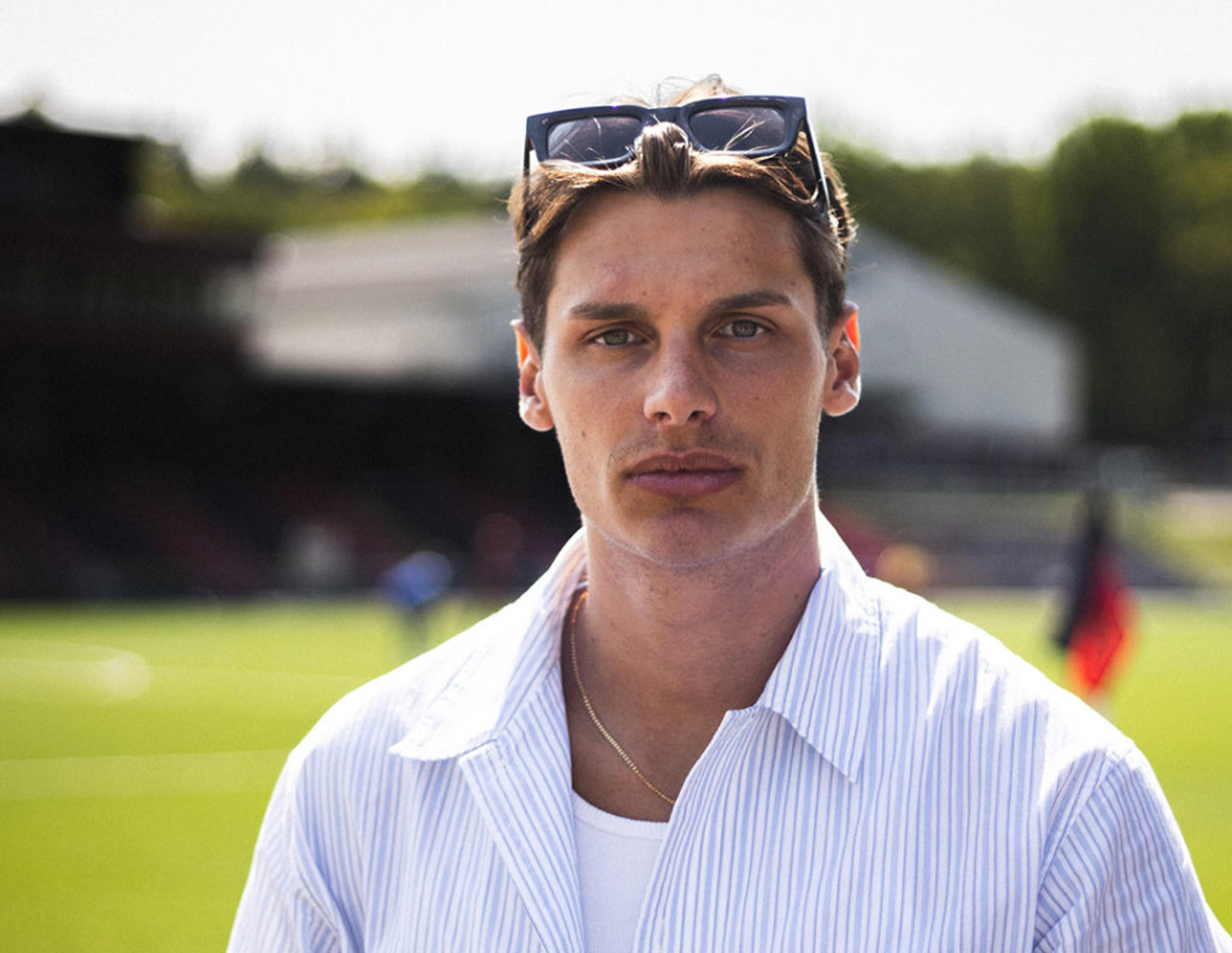 En ung man med solglasögon i håret bär en ljusblå, randig skjorta över en vit t-shirt. Bakgrunden visar en grön fotbollsplan i solljus.