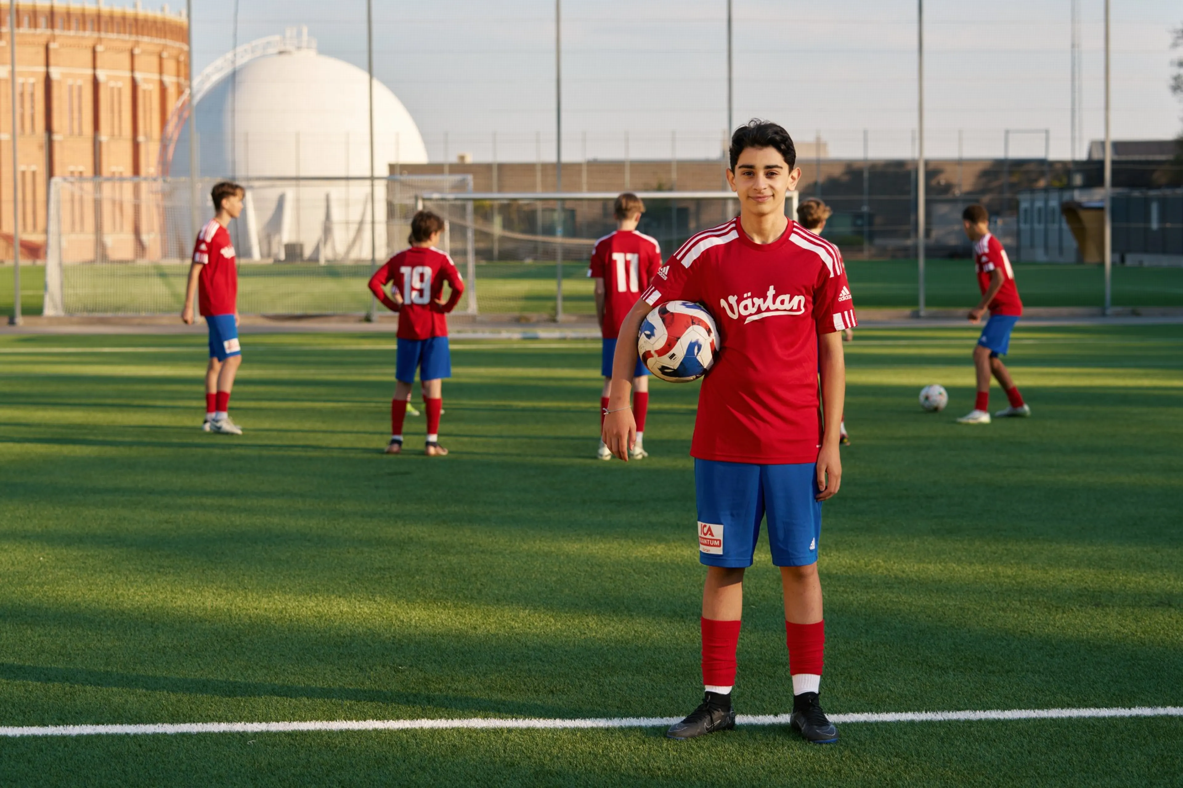 Ett kille i fotbollskläder som har en fotboll under armen och sitt lag i bakgrunden